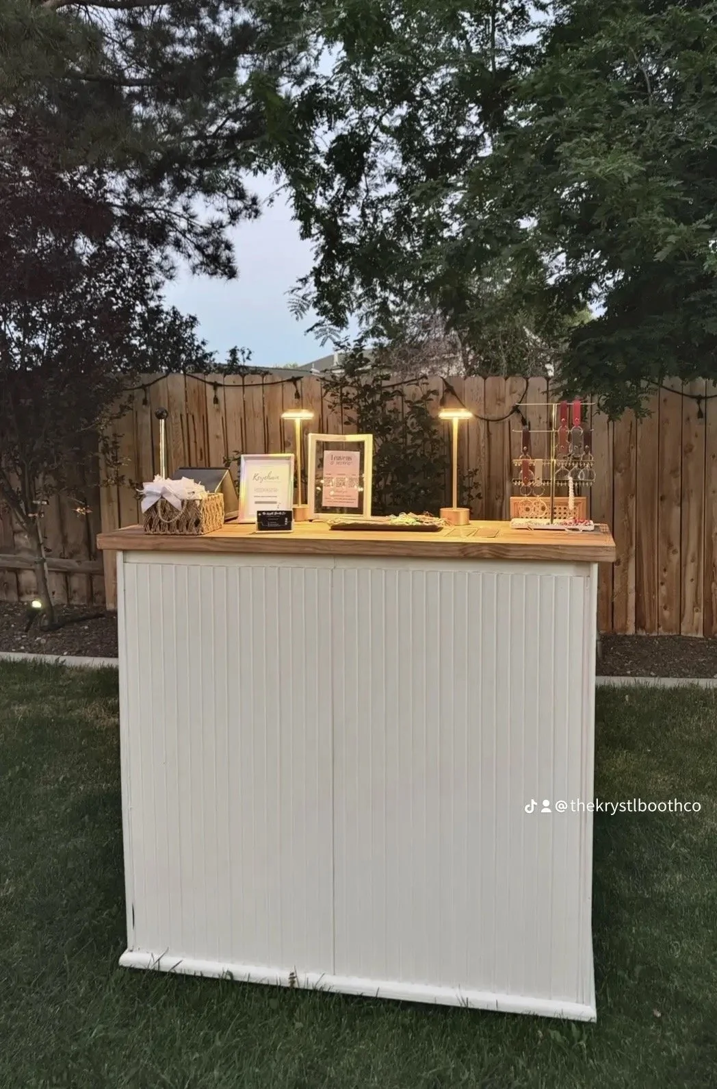 A decorated outdoor event setup with a white and wood-colored bar, framed signs, basket with a bow, and jewelry display stands, illuminated by string lights and small lamps, against a background of trees and a wooden fence.