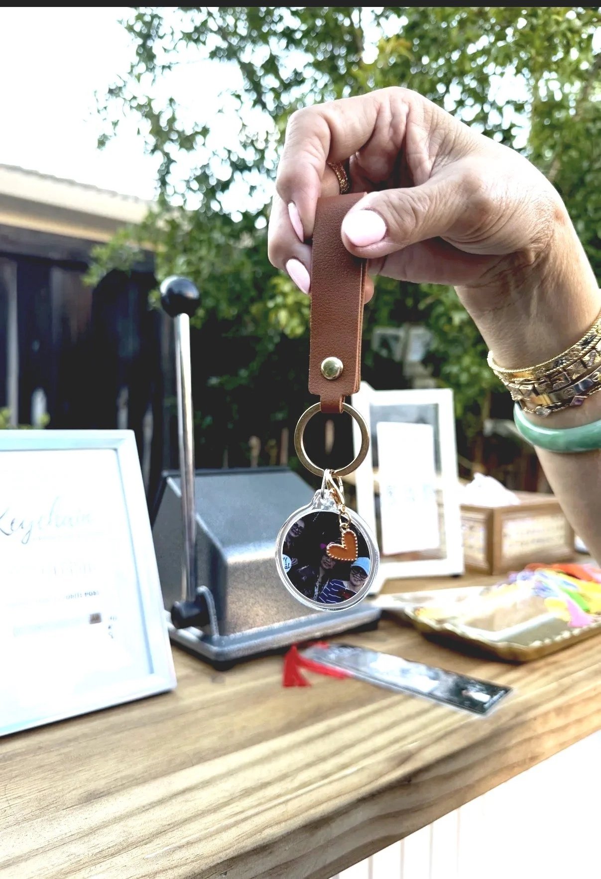Hand holding a brown leather keychain with a circular photo and a small orange heart. The background shows a table with a sign, a kitchen scale, and a tray with colorful items, outdoors with trees and a house.