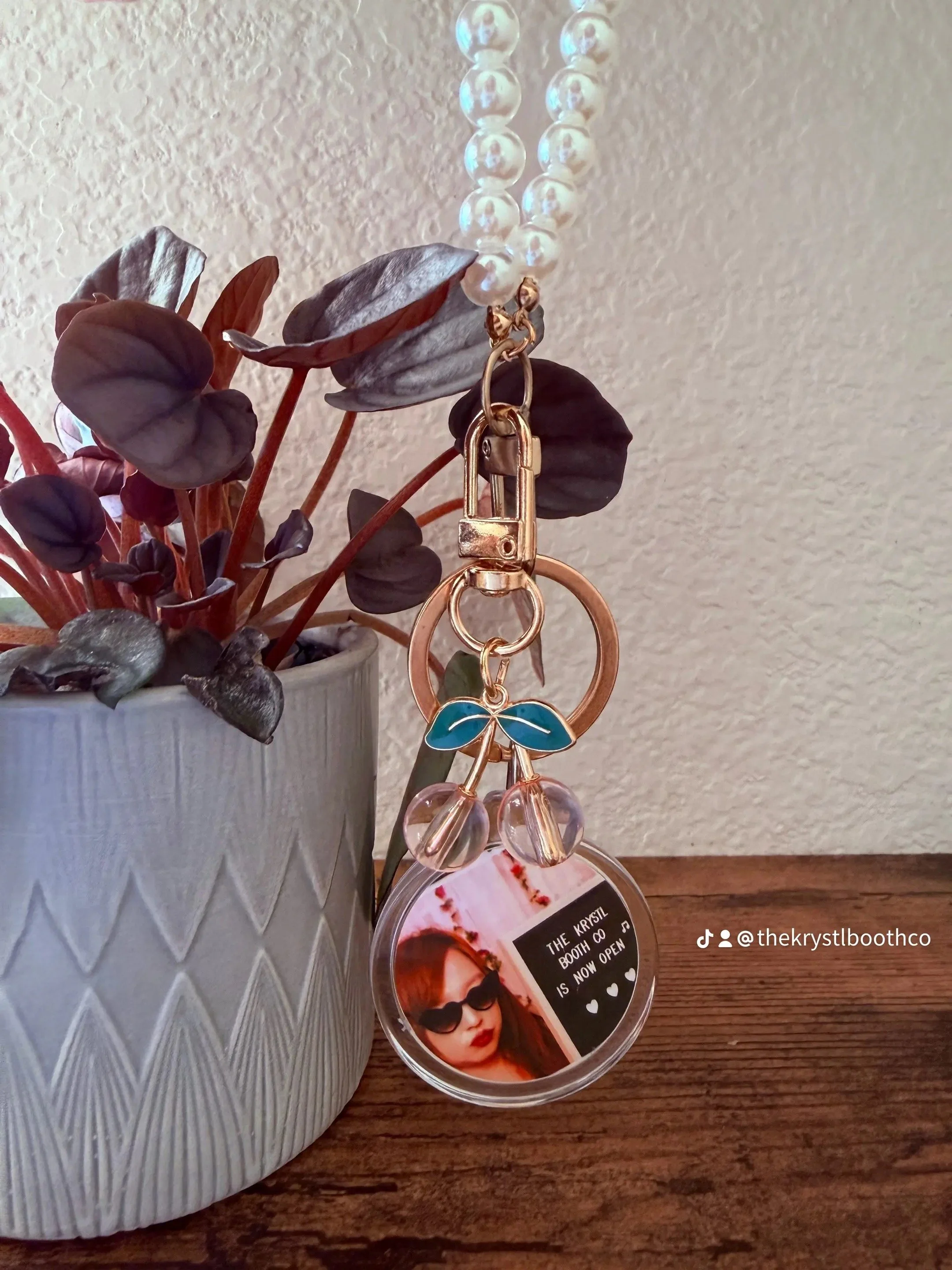 A decorative plant in a textured white pot with dark leaves, a pearl bracelet hanging from a keychain with a blue and gold butterfly charm, and a circular photo of a woman with sunglasses and red lipstick with a message about a photo booth being open.