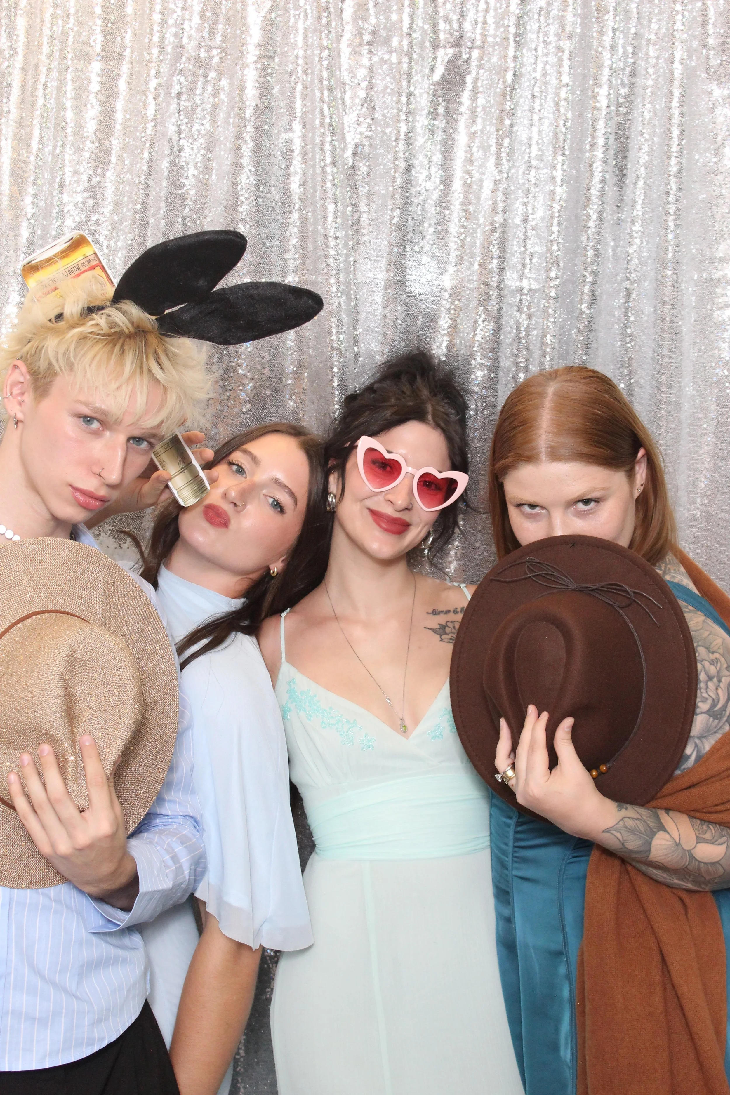 Four women posing in front of a silver, glittery backdrop, wearing various accessories including hats, sunglasses, and bunny ears.
