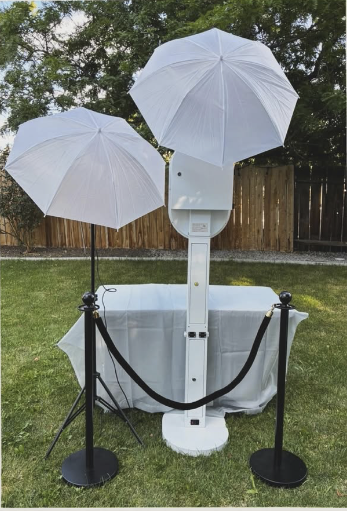 A photo of a photo booth setup in a backyard with two white umbrellas, a table covered with a white cloth, a white photo booth machine, and black stanchions with a retractable belt.