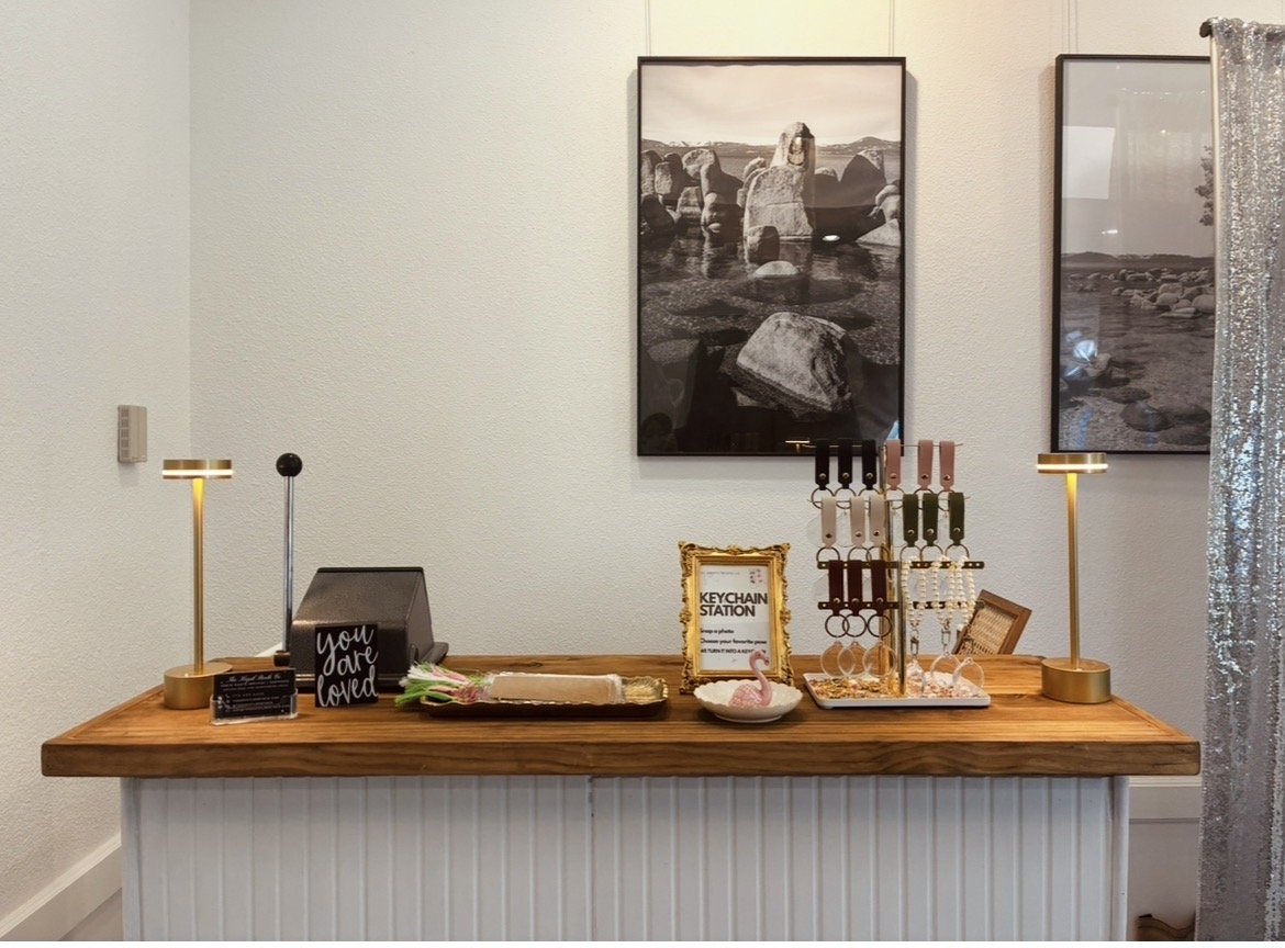 Photo of an indoor setup with a wooden table decorated with lamps, framed signs, and keychains. Two black-and-white landscape art prints hang on the wall above the table, which is covered with a white skirt. Part of a shiny curtain is visible on the right side.