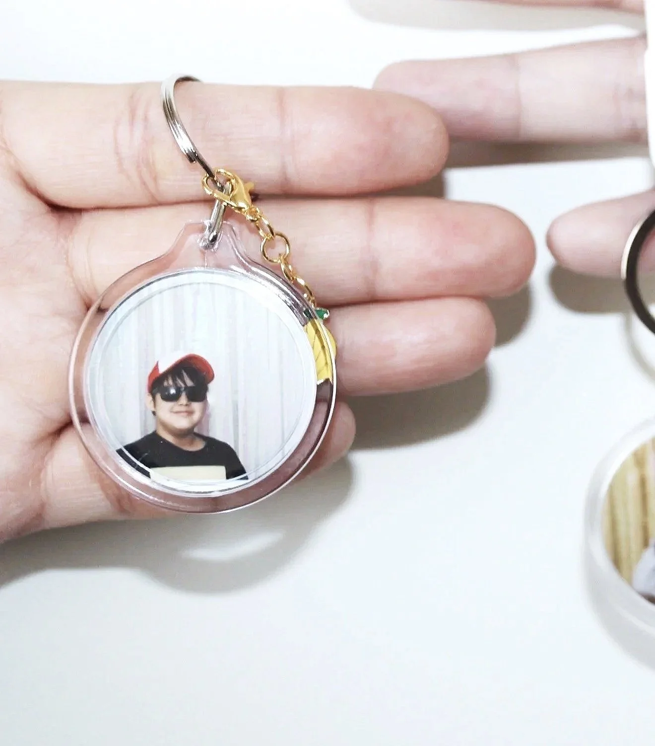 Keychain with a round photo of a young person wearing sunglasses, a black shirt, and a red cap, held by a person's fingers.
