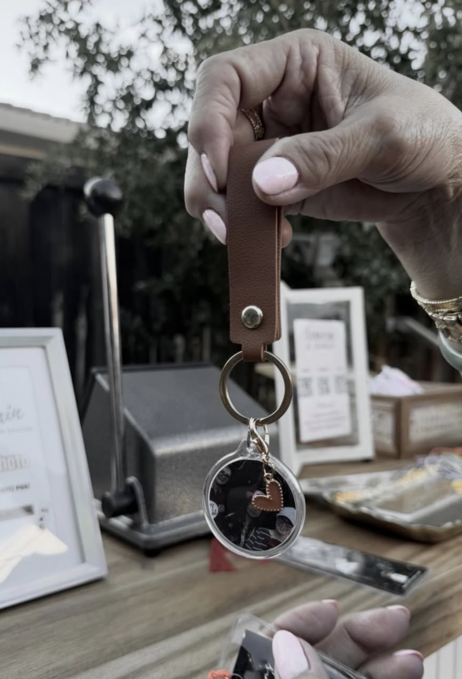 Person holding a keychain with a round mirror and a heart charm above a wooden table with framed menus and other small items, outdoor setting with trees in the background.