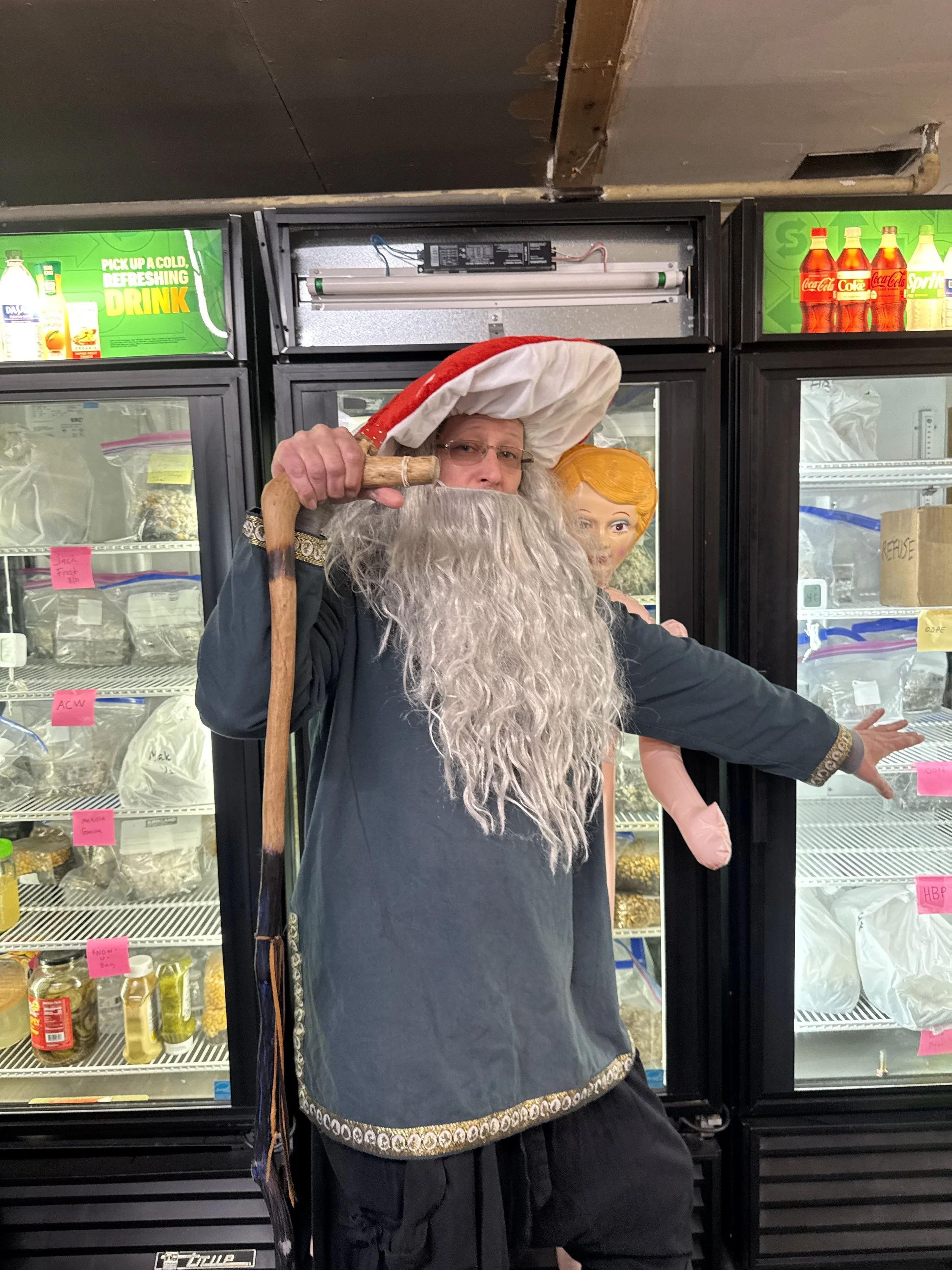 A person dressed as Santa Claus with a long white beard, wearing a red and white Santa hat, standing in front of a grocery store freezer display with various bottles of soft drinks. The person is holding a wooden staff and has one arm extended to the side.