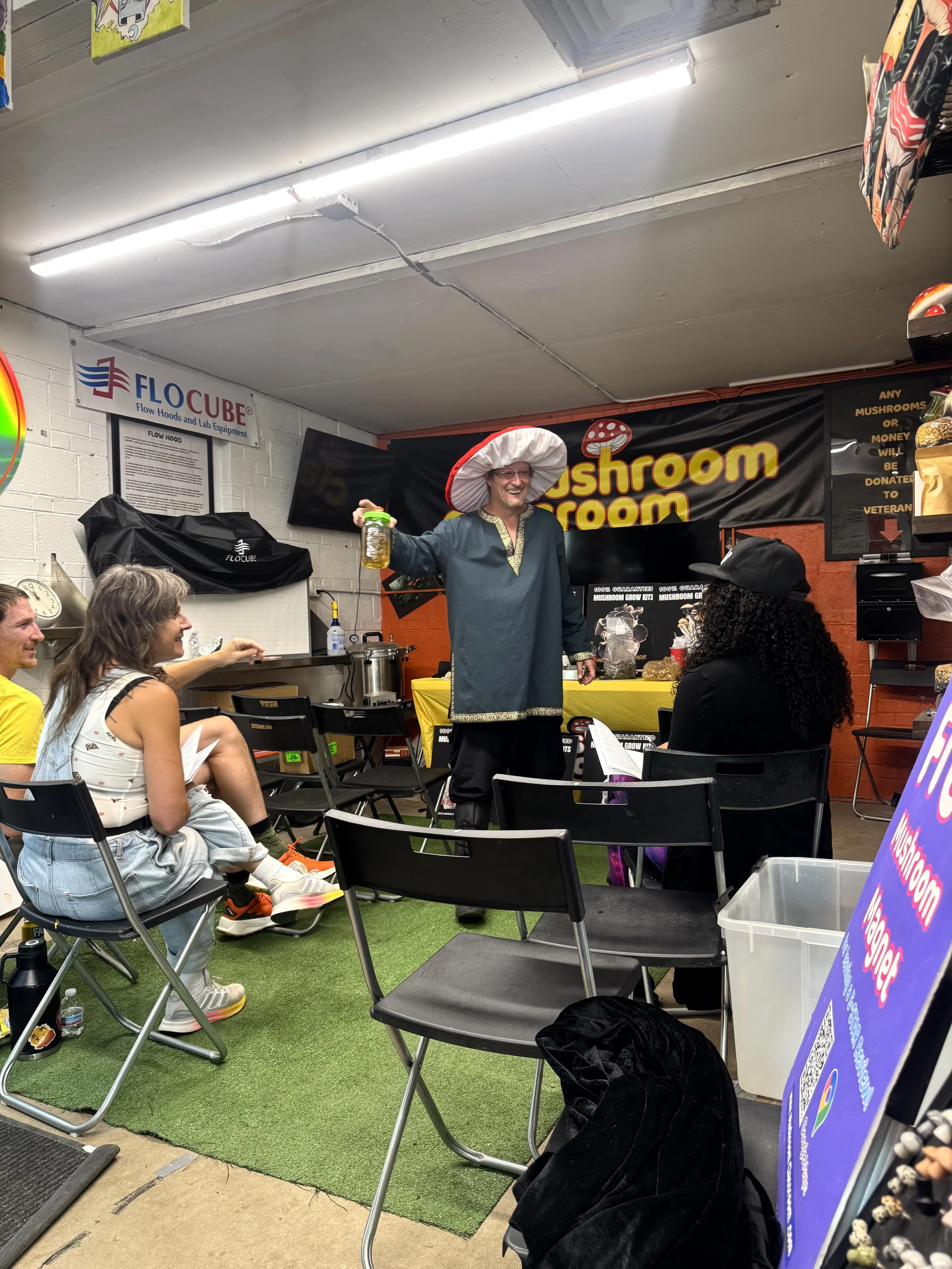 A man dressed in a medieval or fantasy costume with a large mushroom-shaped hat, standing in front of a small group of people seated in black chairs indoors. The background has various signs and decorations, including a banner that reads 'Mushroom' and a sign for 'FLOCUBE Flow Hoods and Lab Equipment.'