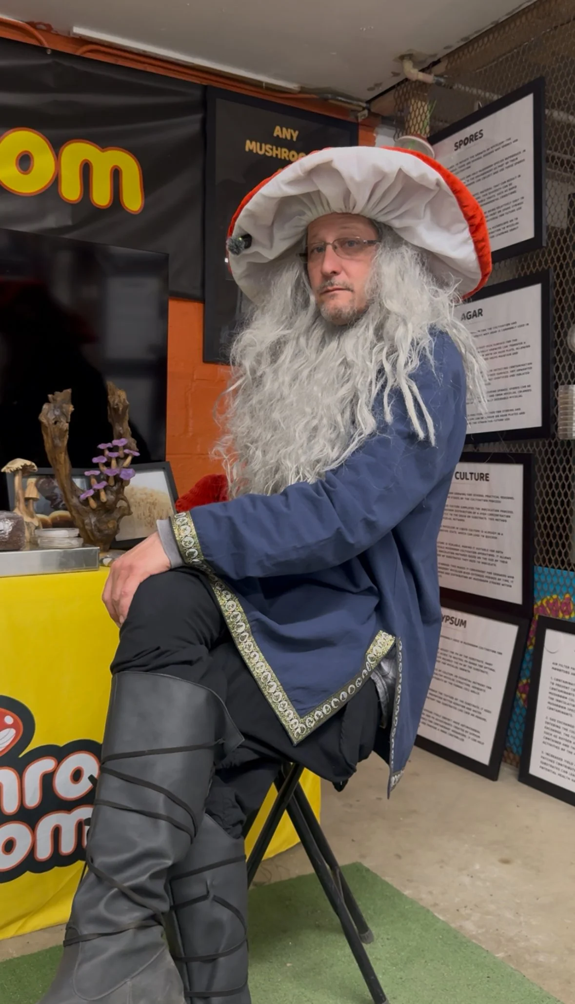 A man dressed as a wizard with a long grey beard, large white and red wizard hat, and medieval-style clothing, seated indoors in front of informational posters.