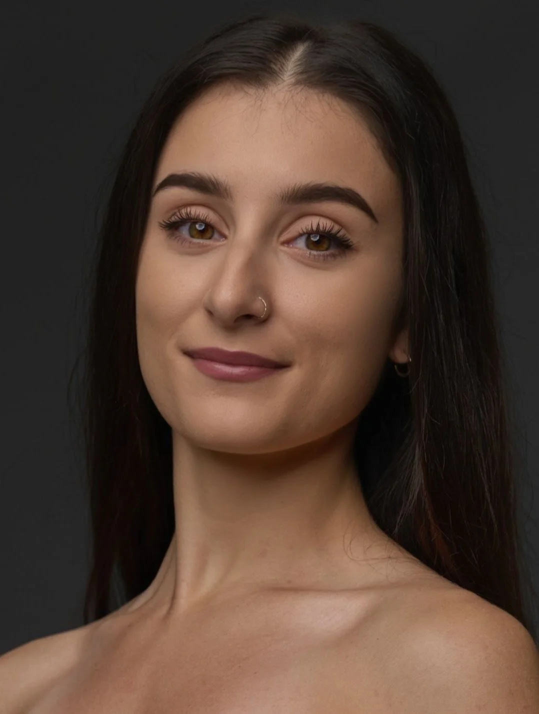 Close-up portrait of a young woman with brown hair, brown eyes, a nose piercing, and light makeup, smiling against a dark background.