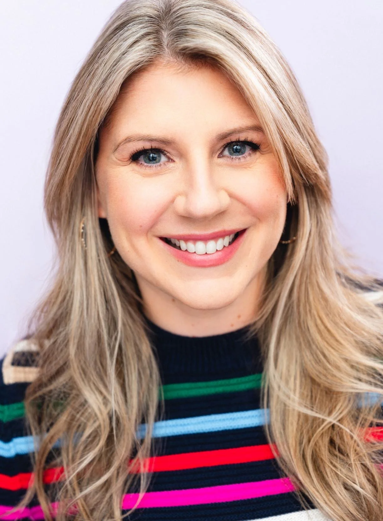A woman with blonde hair and blue eyes smiling, wearing a colorful striped sweater, in front of a plain light background.