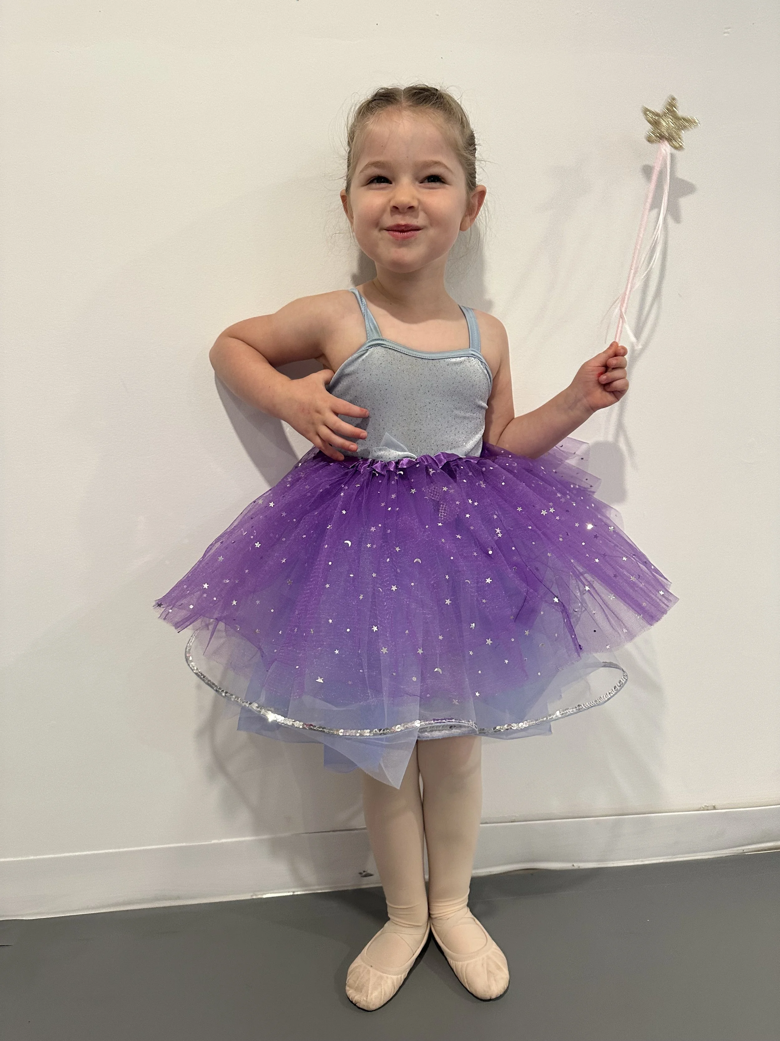 A young girl dressed as a ballerina in a purple tutu, silver leotard, ballet slippers, holding a star-shaped wand, standing against a plain white wall.