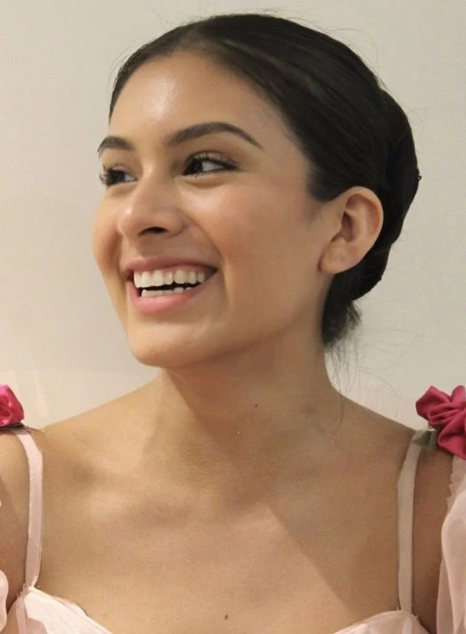 A woman smiling and looking to the left, with dark hair styled in a braided updo, wearing a pink top with pink shoulder accents, against a plain light background.
