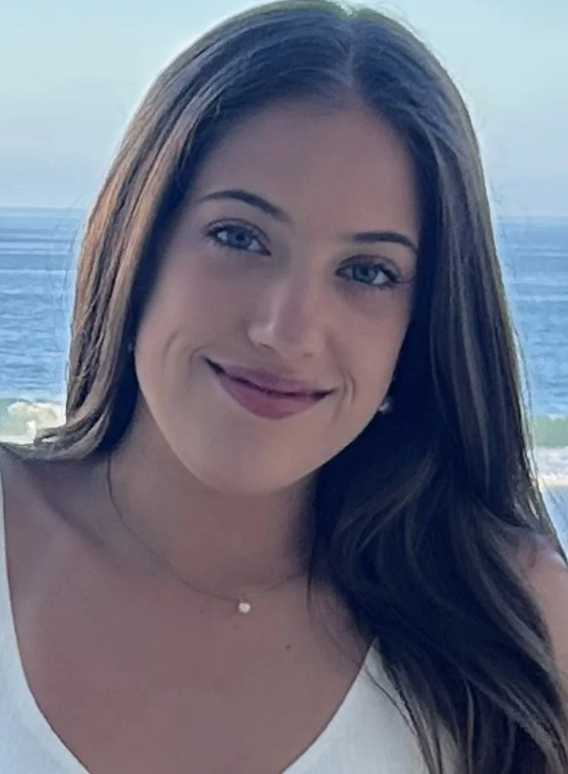 A young woman with long brown hair smiling at the camera with the ocean and sky in the background.