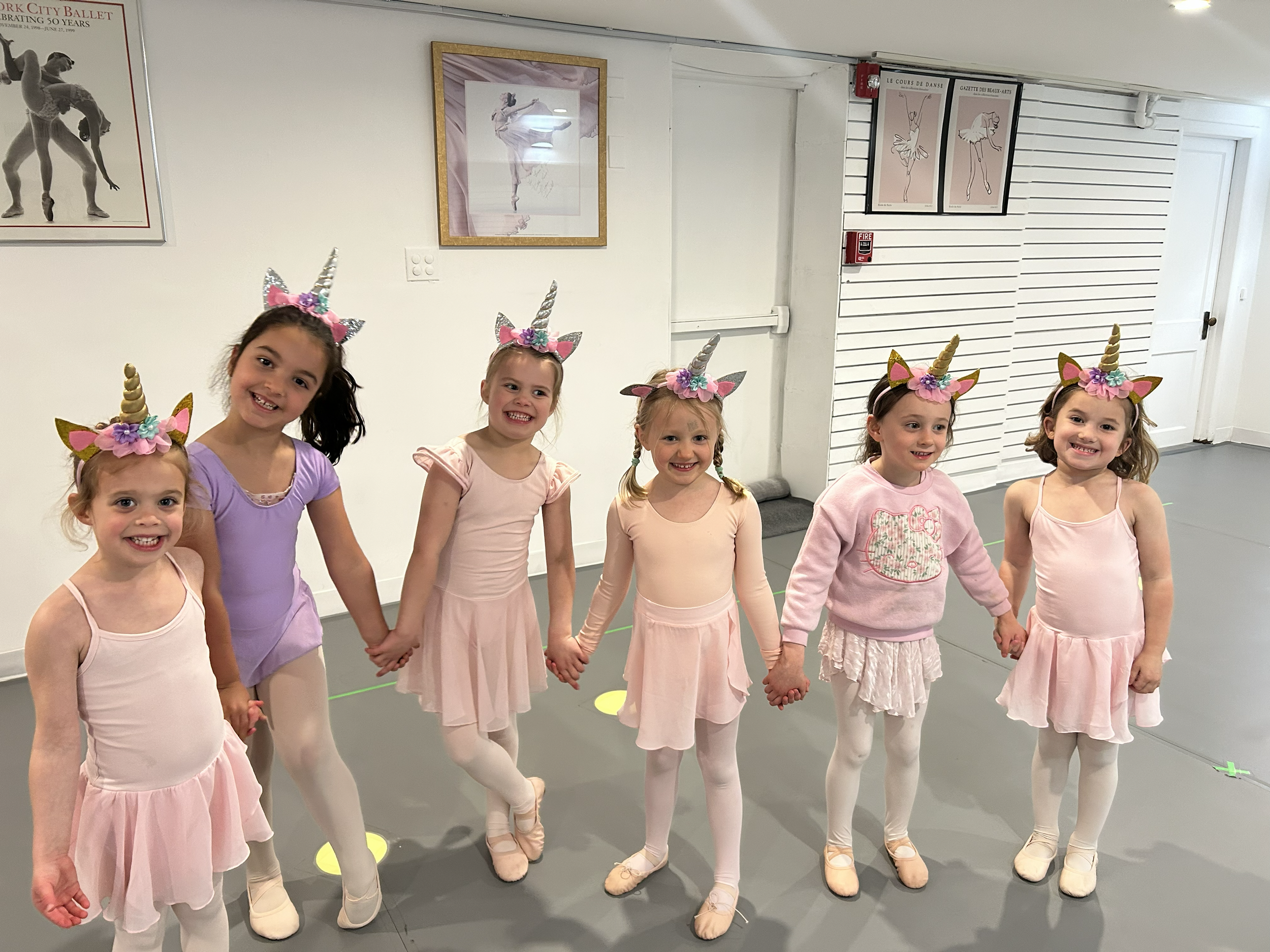 Six young girls wearing ballet costumes with unicorn horns and ears, standing in a dance studio holding hands in a line, smiling. The studio has framed dance posters on white walls and a door in the background.