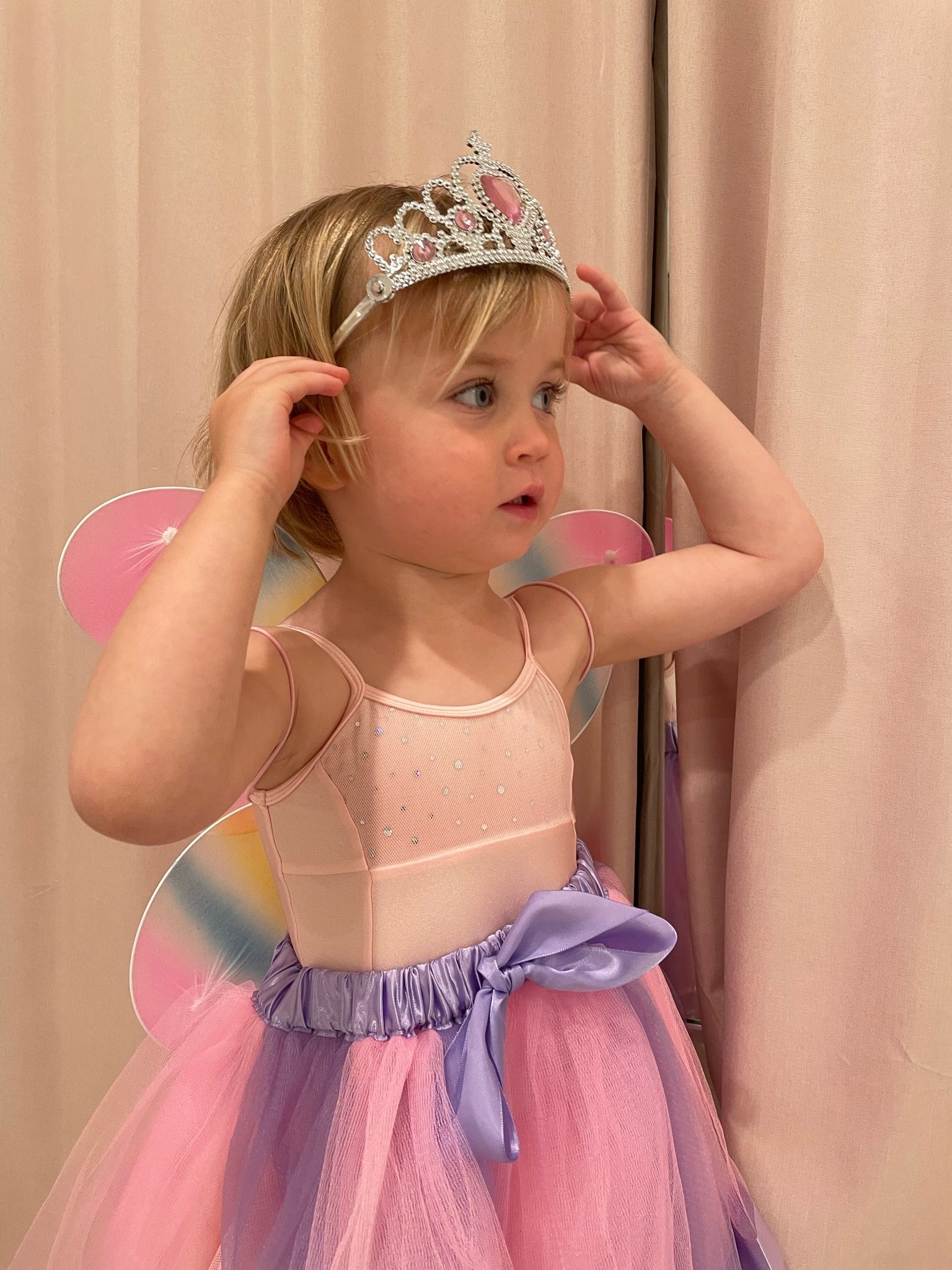 Child dressed as a fairy princess, wearing a pink and purple tutu, a silver tiara, and pink fairy wings, standing in front of a light pink curtain.