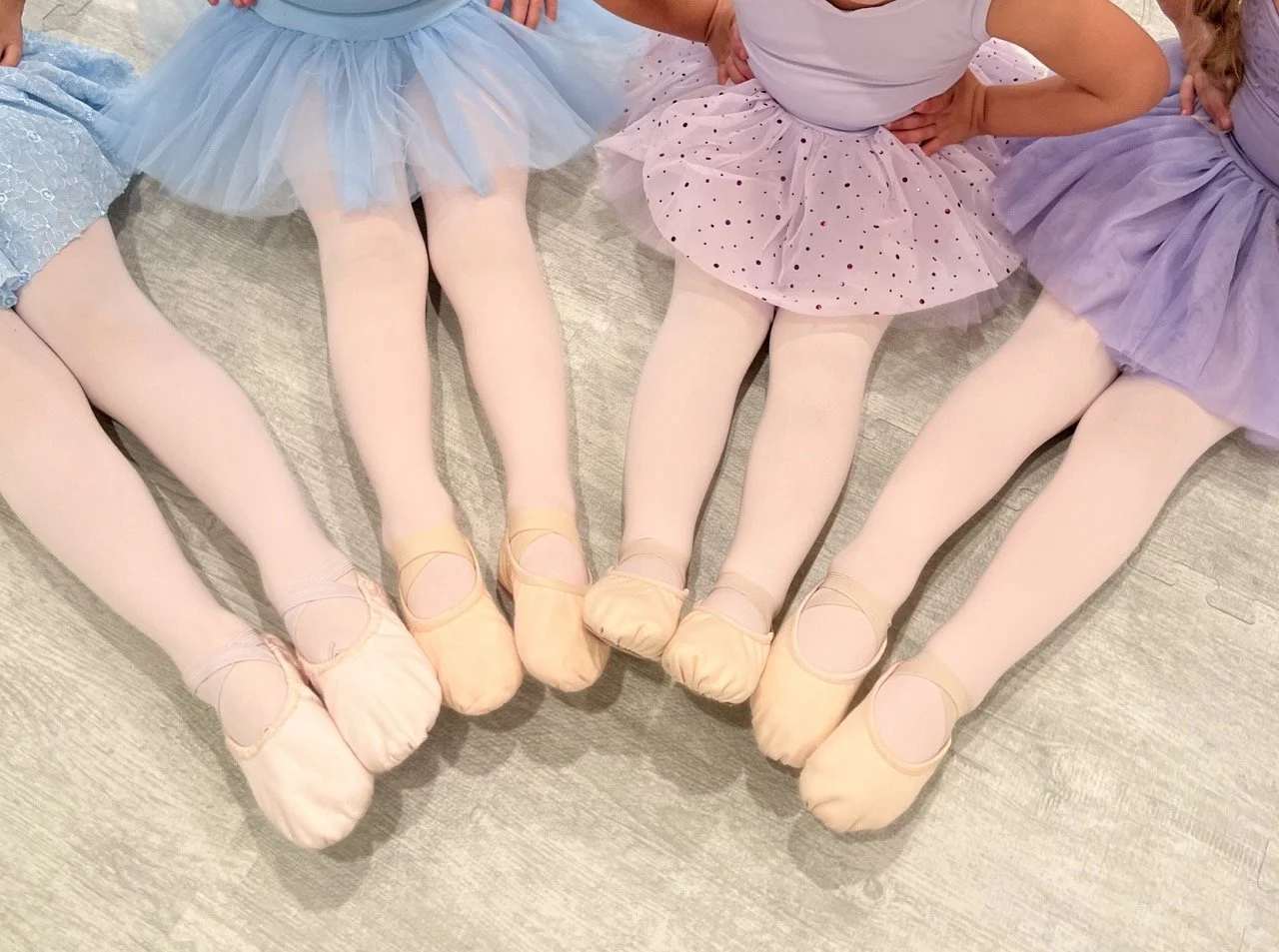 Four young girls dressed in ballet tutus and tights, sitting on the floor with their legs extended and feet in toe shoes.
