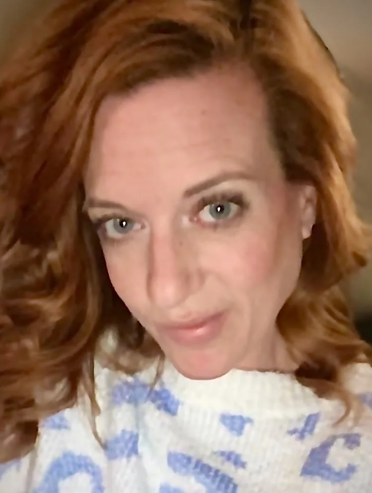 Close-up selfie of a woman with wavy red hair and blue eyes, wearing a white top with blue patterns.