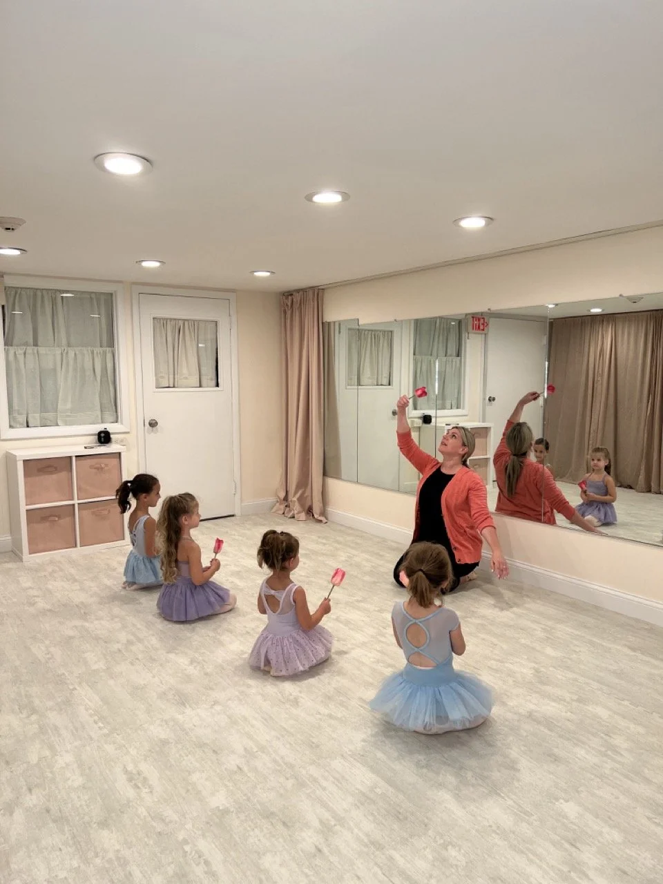 A dance instructor in a coral jacket leading a ballet class for young girls in tutus, sitting on the floor with ballet slippers in a dance studio with a large mirror.