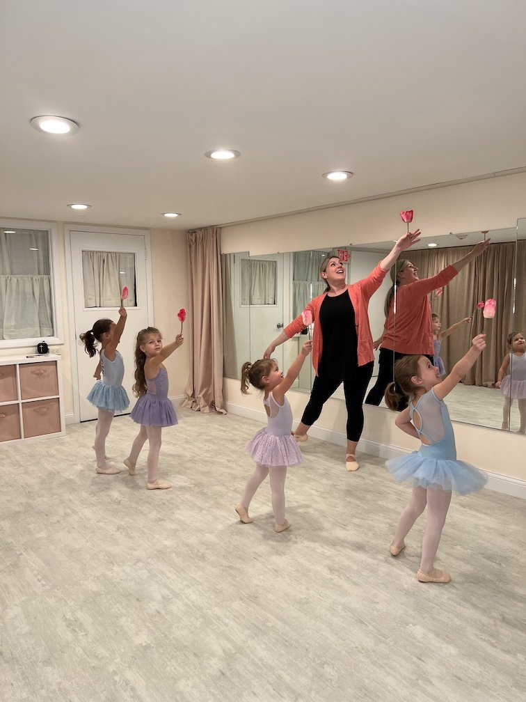 A ballet instructor leading five young girls in a ballet class, all holding pink paper flowers, in a dance studio with a large mirror.