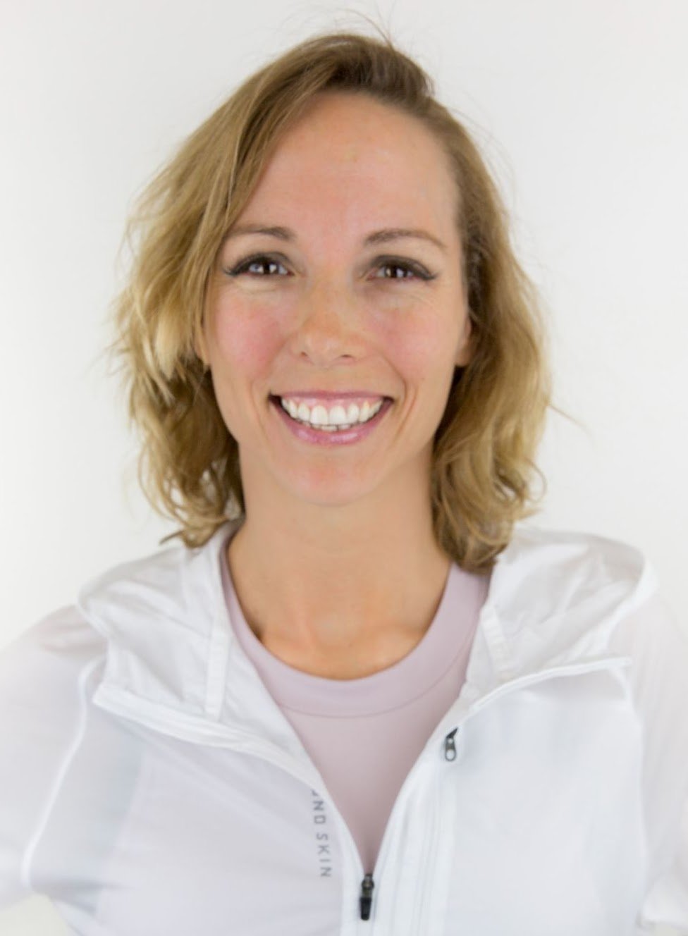A woman with shoulder-length blonde hair smiling, wearing a white jacket and a light pink shirt against a white background.