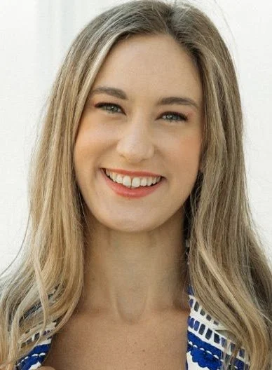 A smiling woman with long blonde hair, wearing a blue and white striped top, standing indoors.