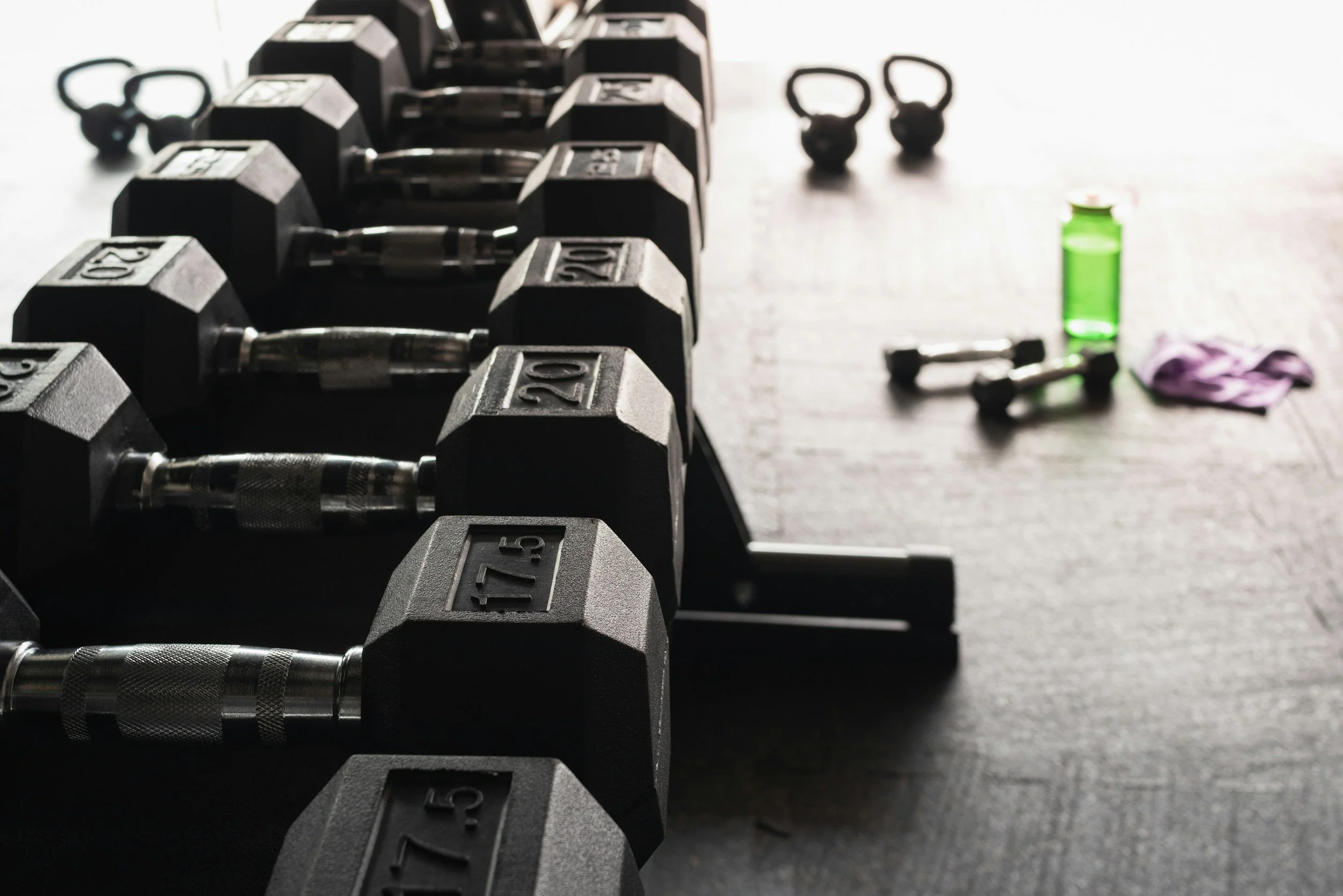 Row of black dumbbells with weight markings, some kettlebells, a green water bottle, and resistance bands on a dark gym floor.