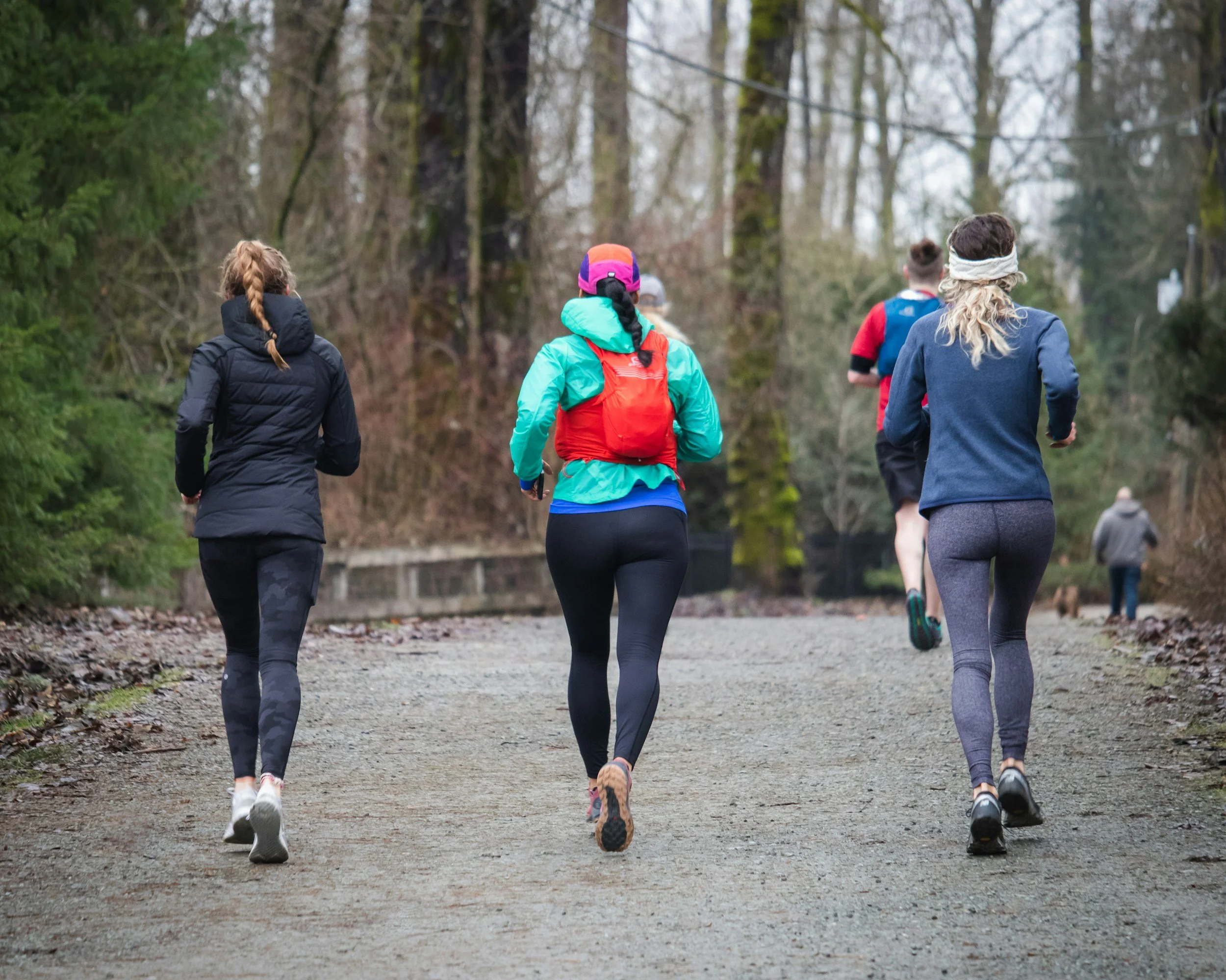 A group of people jogging or running on a gravel path through a forest or wooded area, with trees on both sides, during daytime.