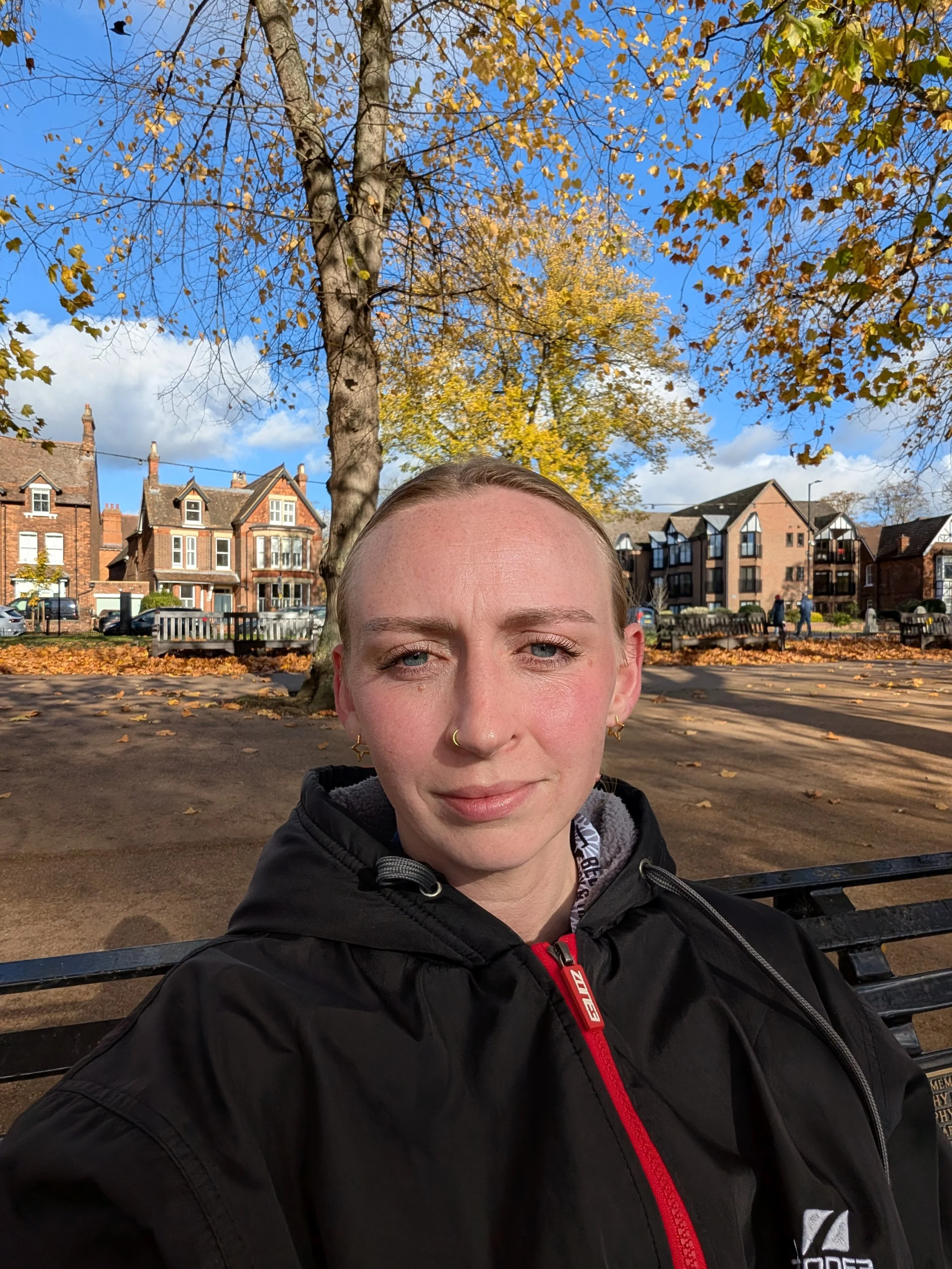 A person with short blonde hair, blue eyes, and light skin sitting on a park bench during daytime, wearing a black jacket with a red zipper. This is a photo of the personal trainer Kathryn.