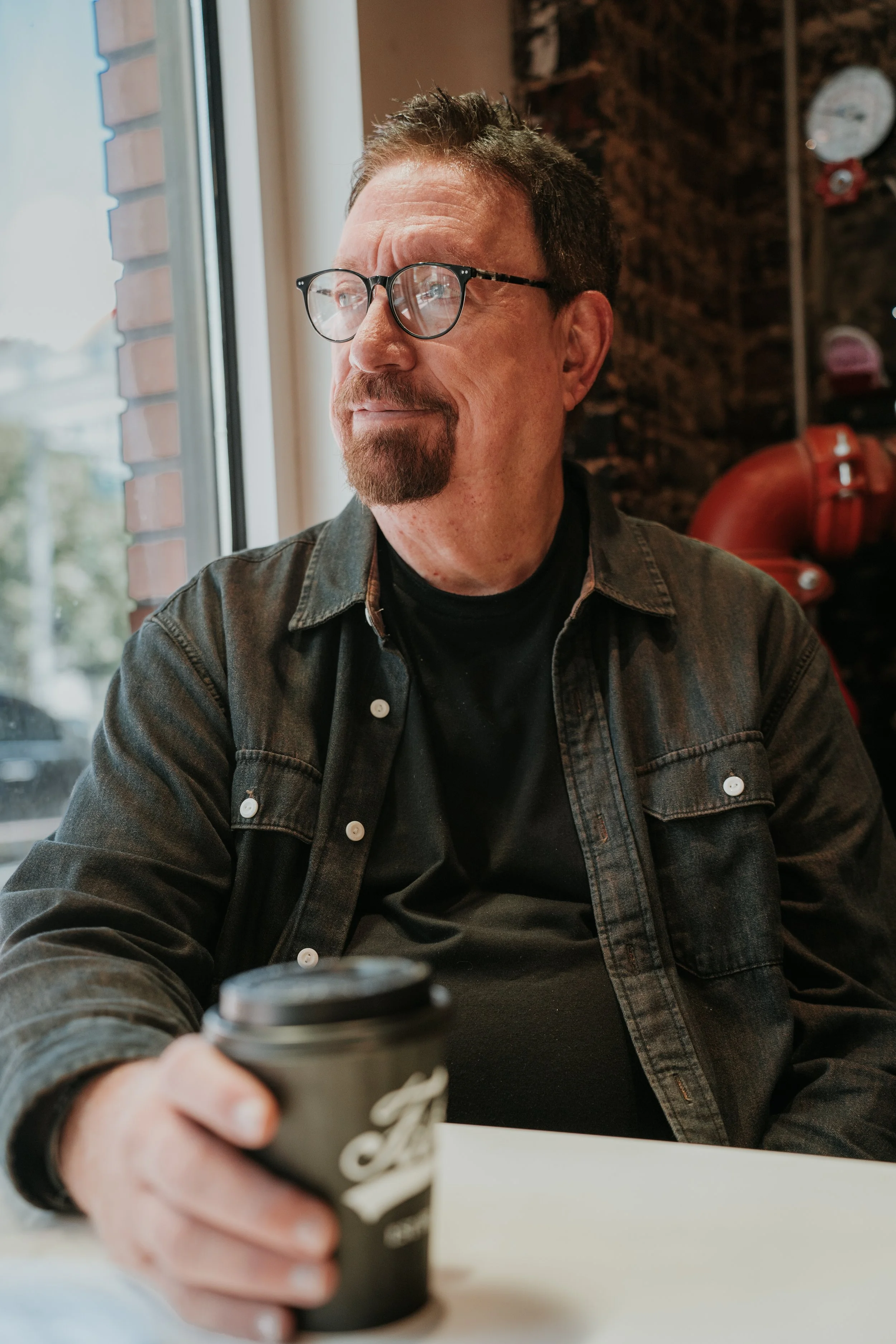 A man with glasses and a goatee is sitting at a table, holding a cup of coffee, with a window and a brick wall in the background.