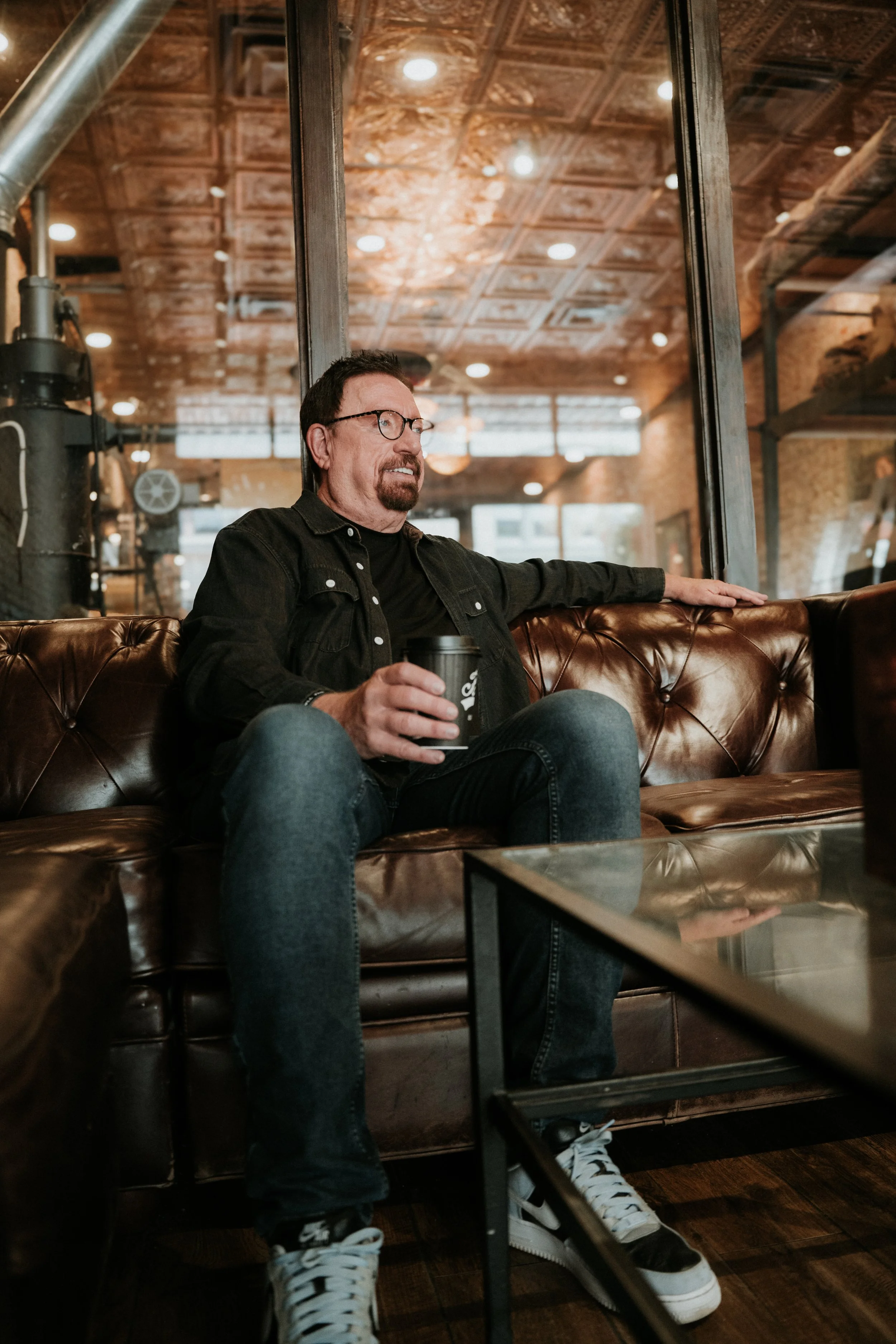 A man with glasses, a beard, and a mustache sits on a leather couch in a coffee shop, holding a coffee cup, wearing a black jacket, black t-shirt, jeans, and white Nike sneakers, with a wooden ceiling and industrial decor visible in the background.