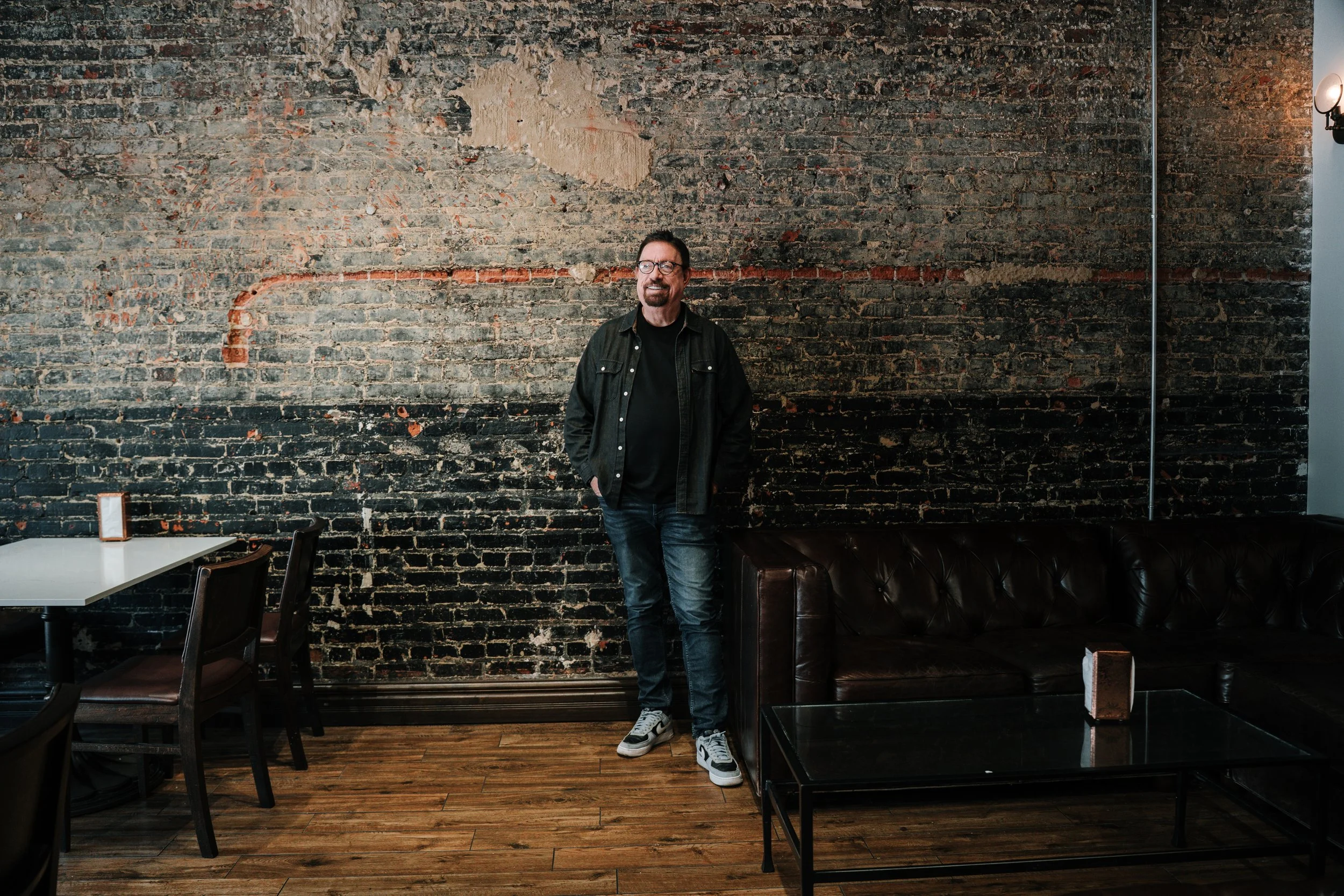 Man with glasses, dark jacket, and sneakers stands in front of an exposed brick wall inside a cafe or restaurant.