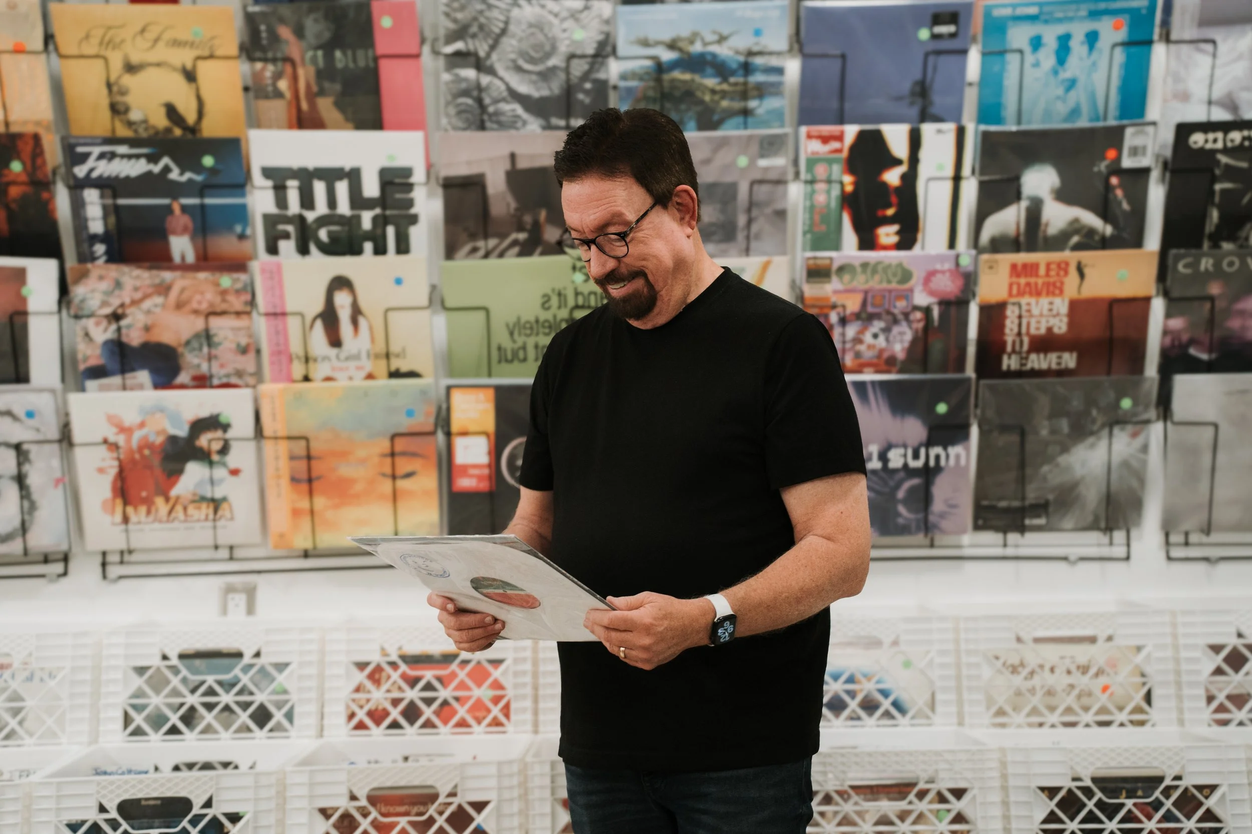 A man with glasses and a beard, wearing a black shirt, is standing in front of a wall filled with vinyl record covers. He is looking down at a vinyl record in his hands and smiling.