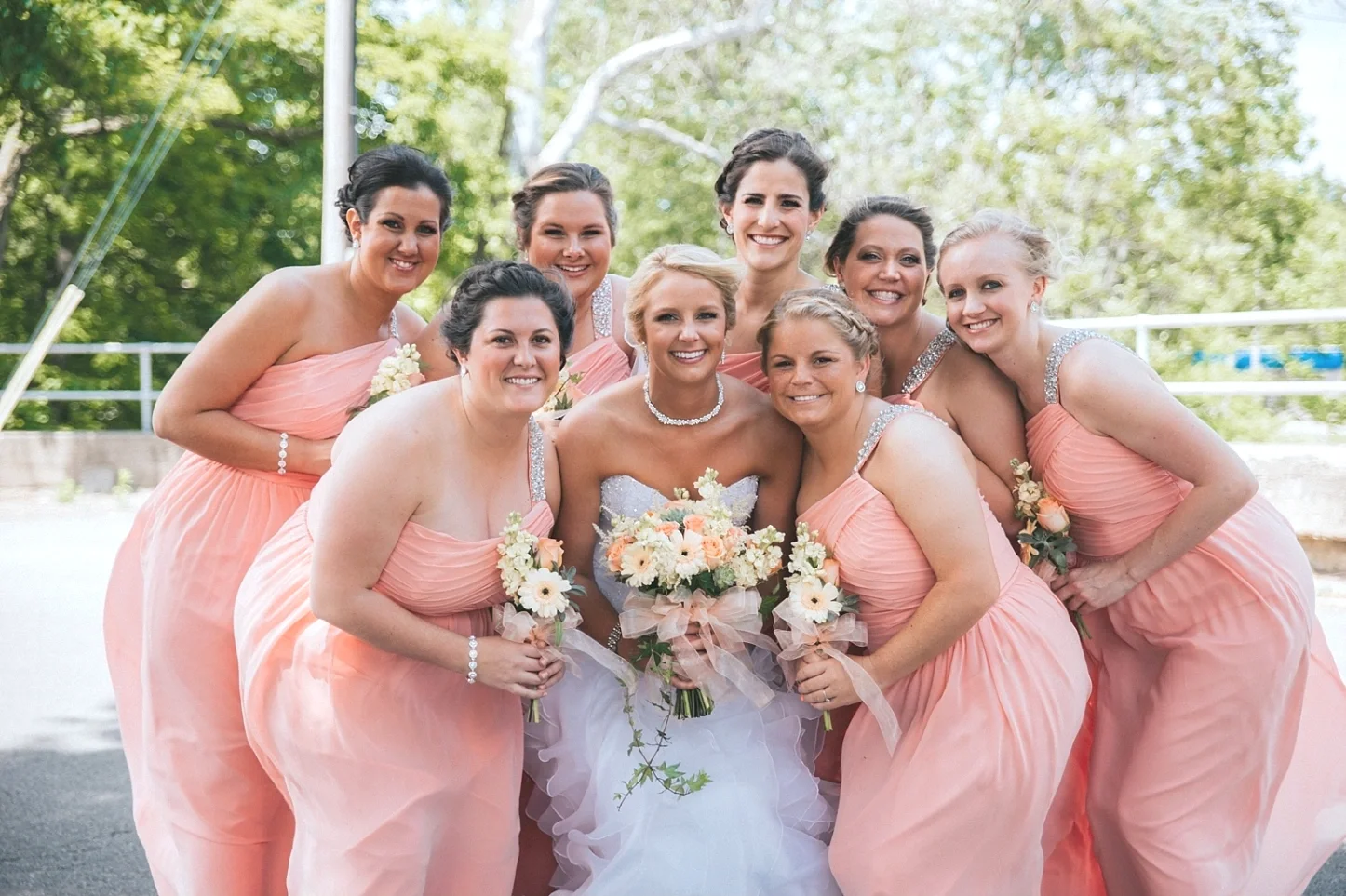A bride and seven bridesmaids in peach dresses, smiling outdoors with trees in the background.