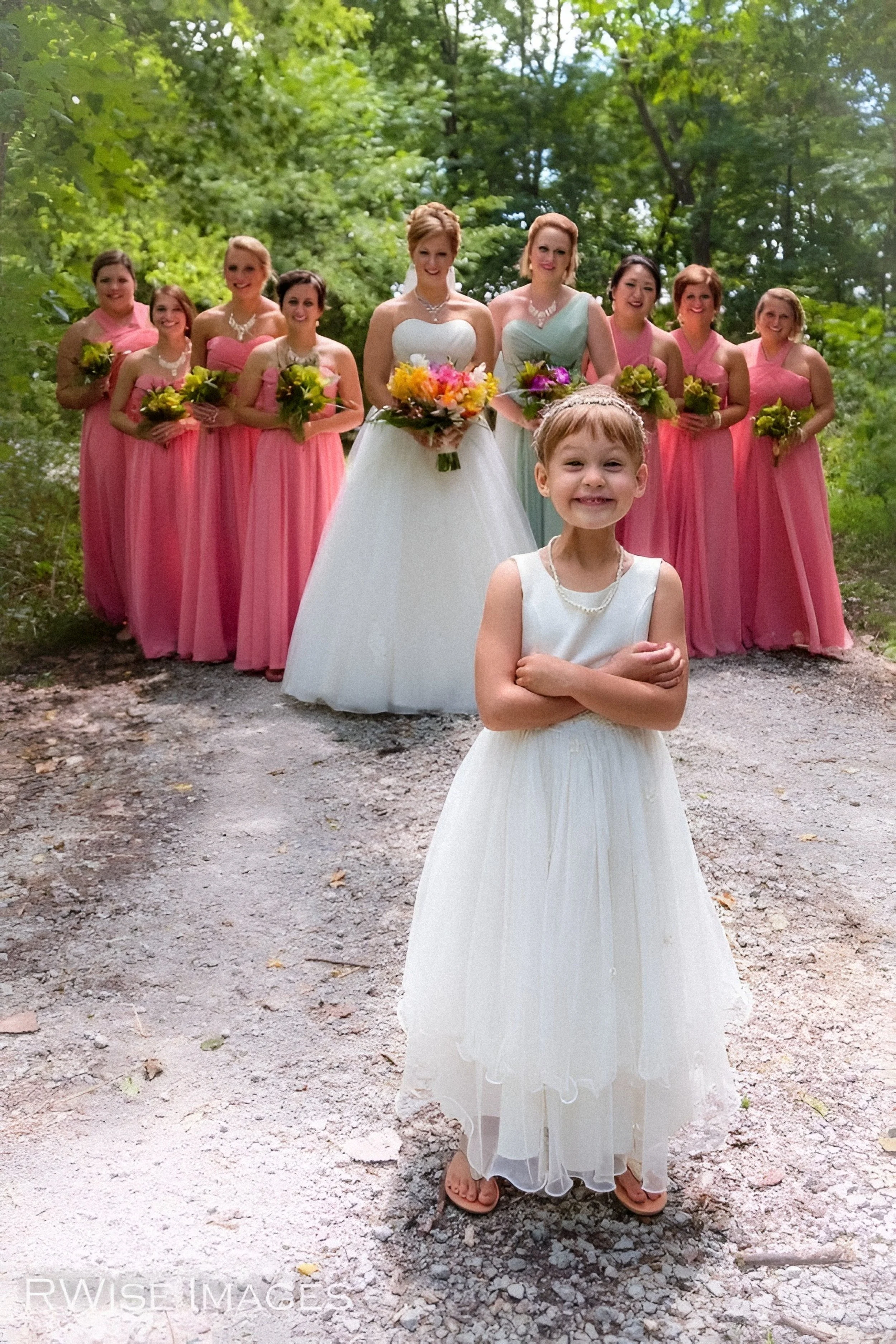 A group of women in pink dresses and one young girl in a white dress, standing outdoors on a dirt path surrounded by green trees, holding bouquets of flowers, with the woman in a white wedding gown at the center of the group.