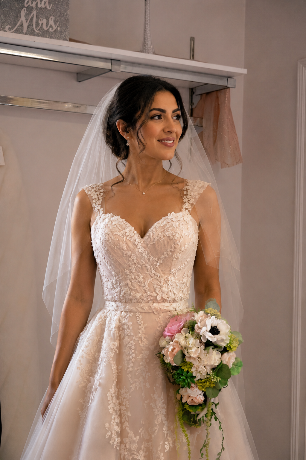 A woman in a wedding dress holding a bouquet, standing indoors with a neutral-colored wall and decorations in the background.