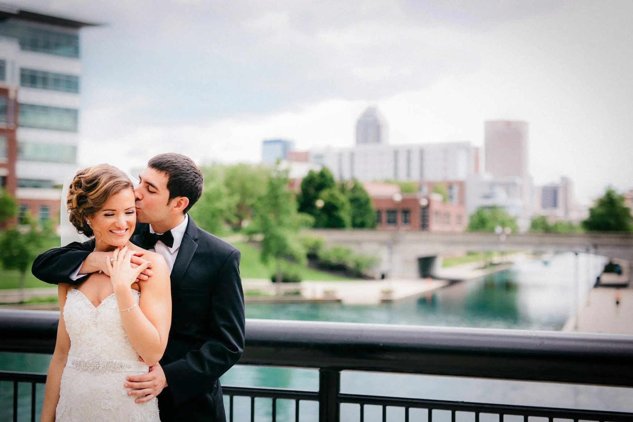A happy bride and groom share a moment of affection outdoors on their wedding day, with the groom kissing the bride on her temple, as she smiles warmly, surrounded by urban buildings and greenery.