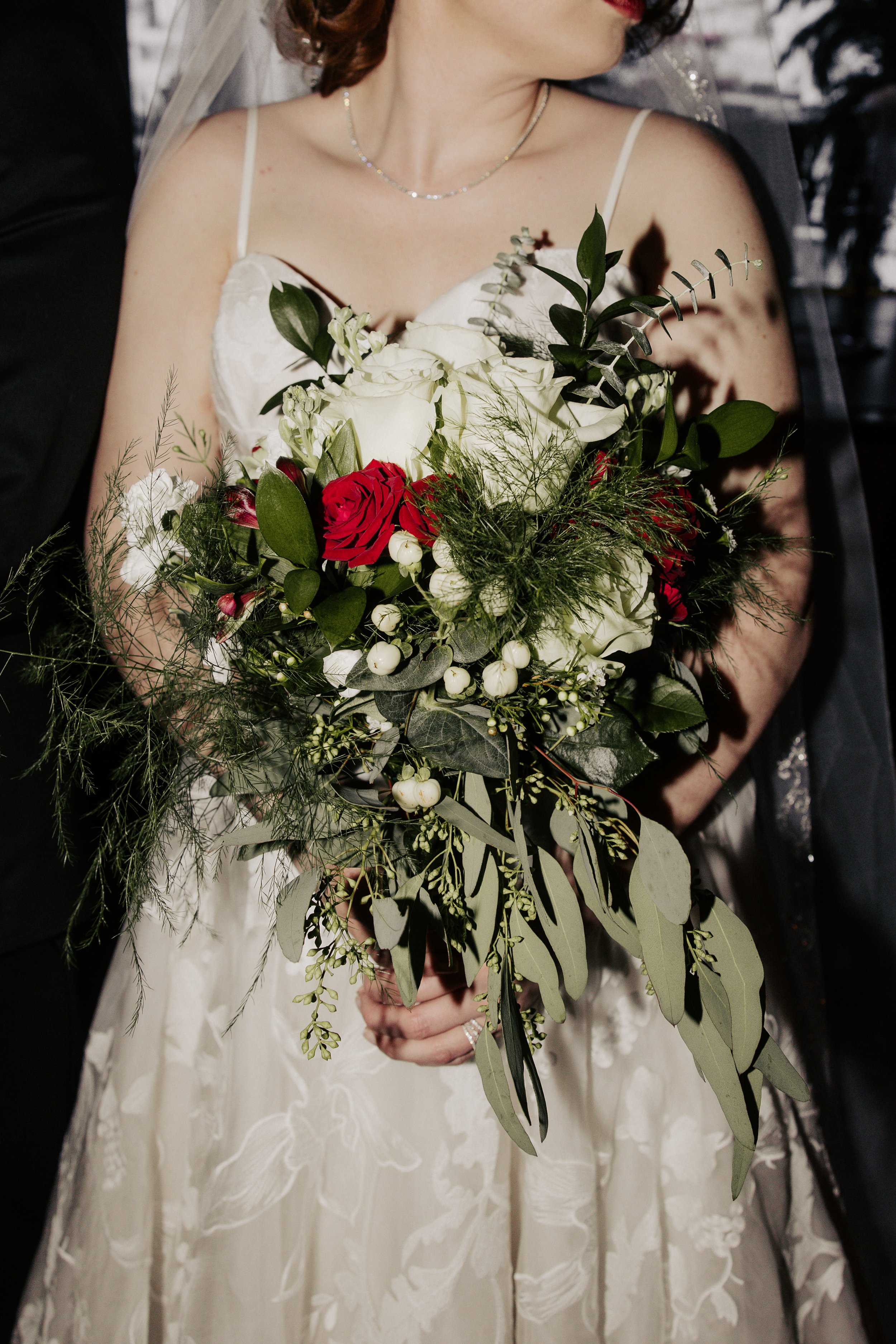 A bride holding a large bouquet of white, red, and green flowers and foliage in front of her white dress.