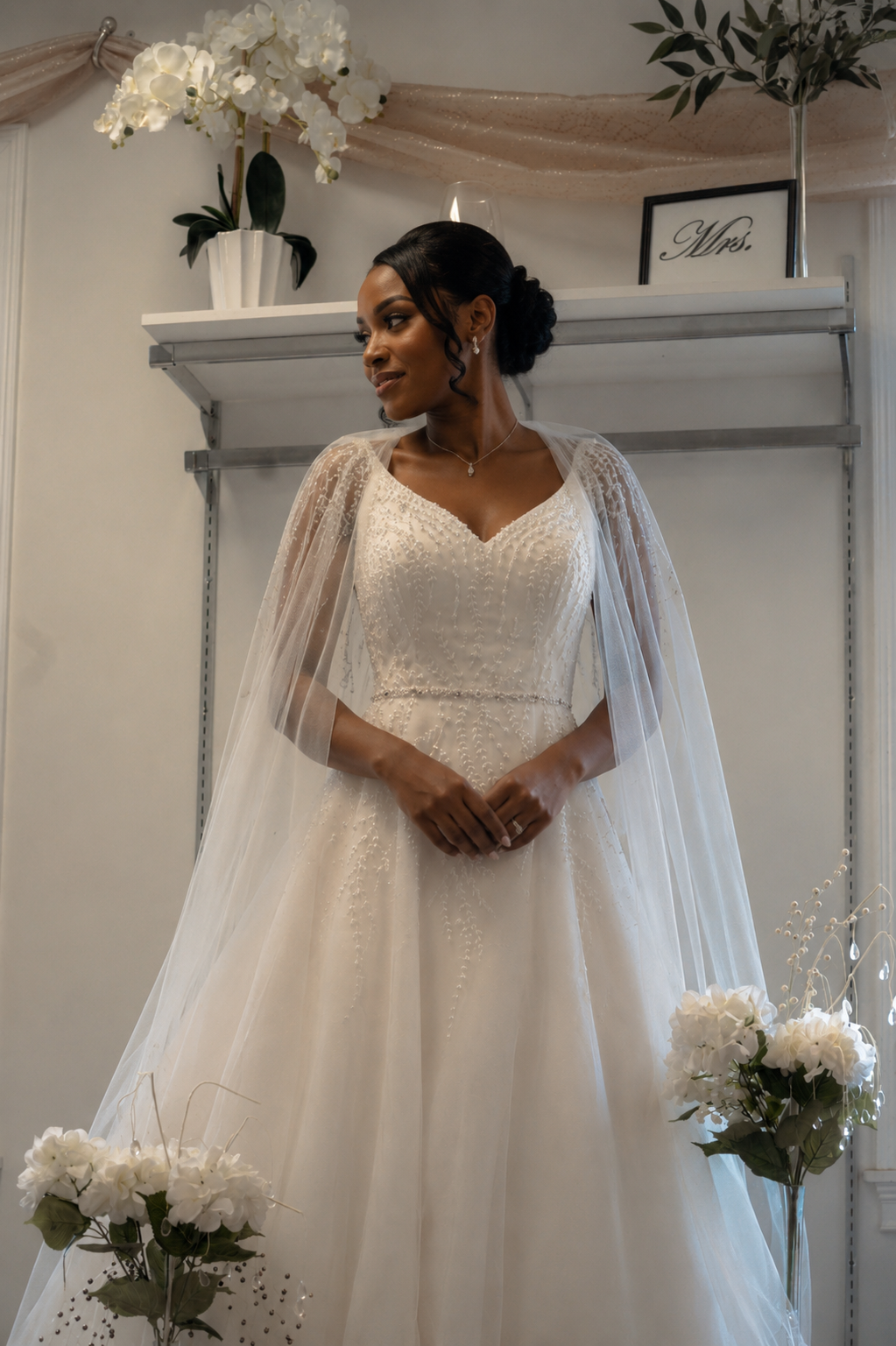 A woman in a wedding dress standing indoors, with floral decorations and a sign that says "Mrs." in the background.