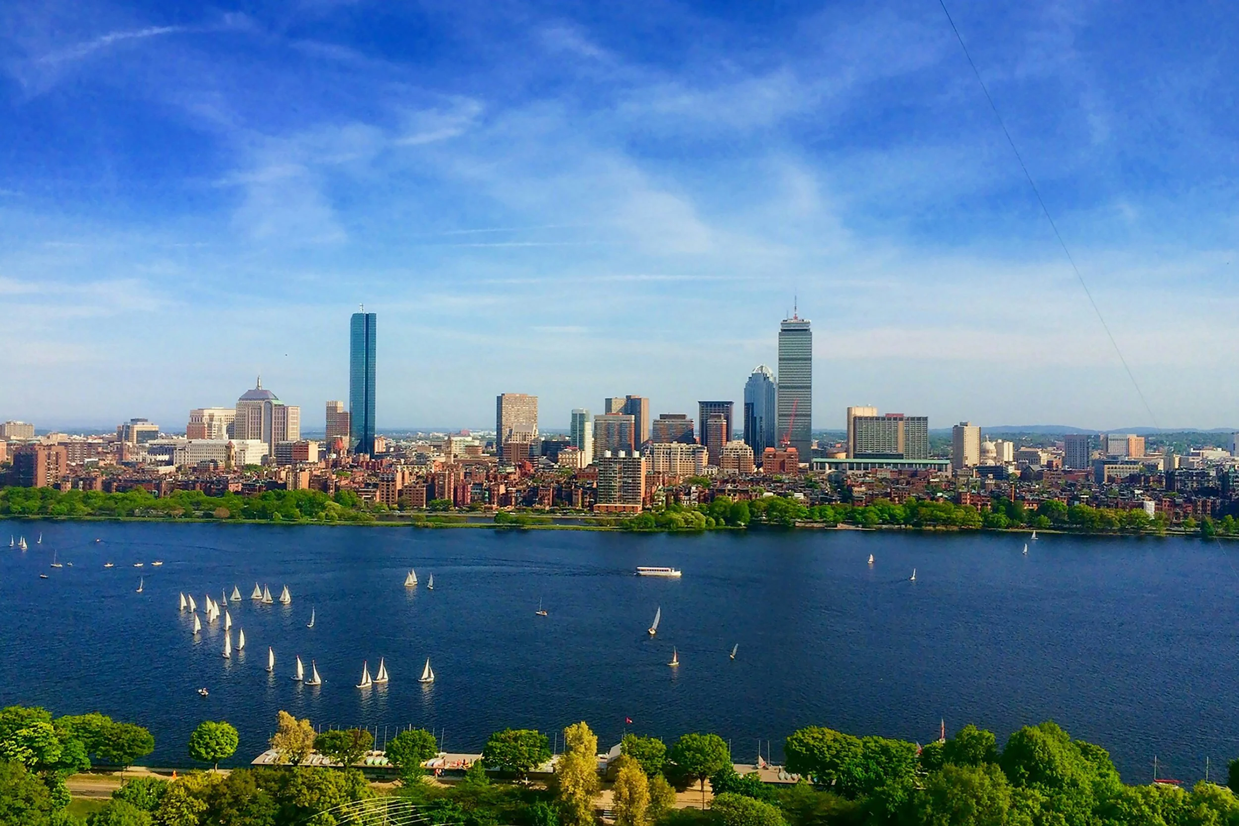 A city skyline with tall buildings, a river in the foreground with sailboats, and green trees at the bottom of the image under a blue sky with some clouds.