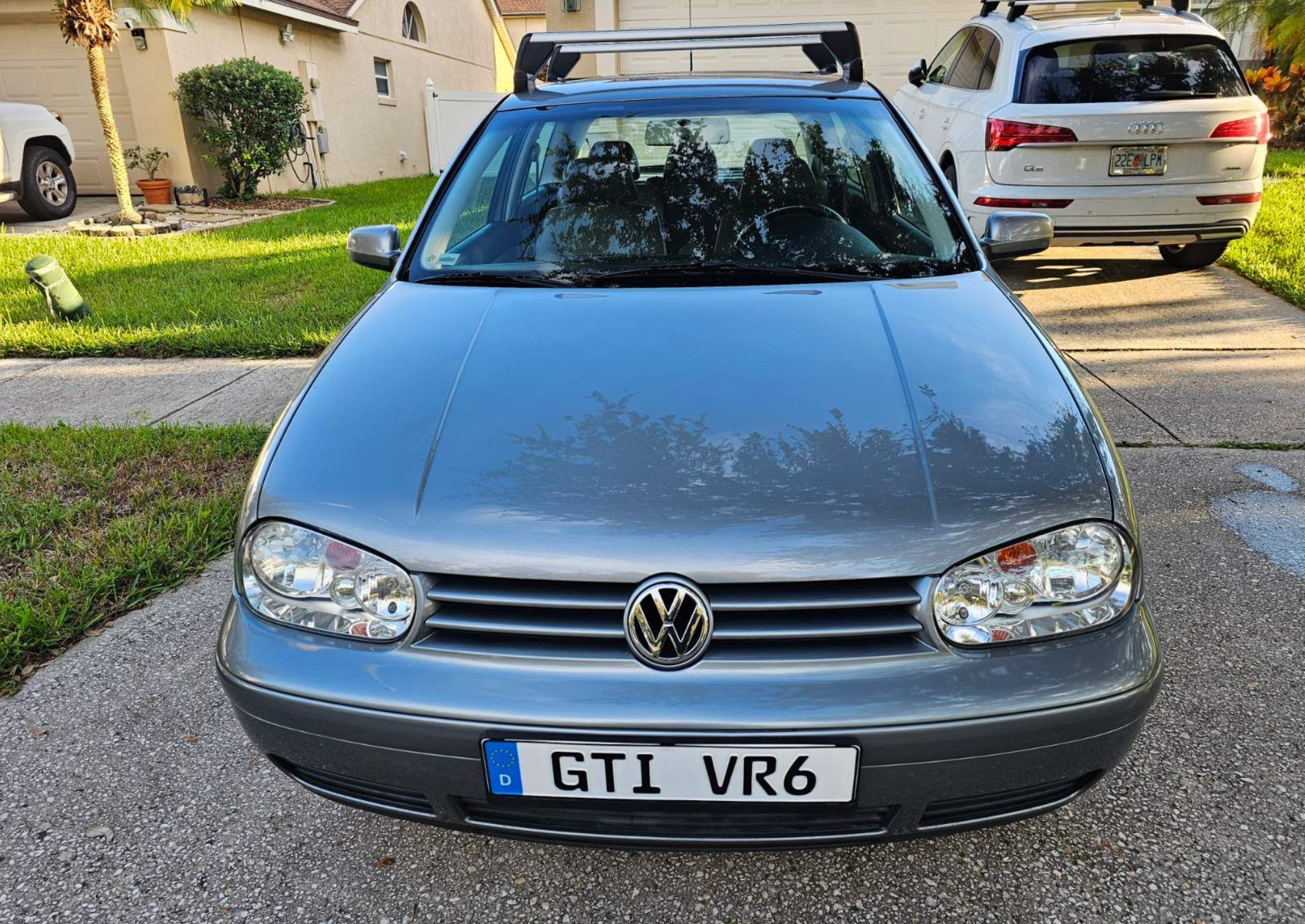 A silver Volkswagen Golf parked in a residential driveway facing the viewer, and part of a house, grass, and trees visible in the background.