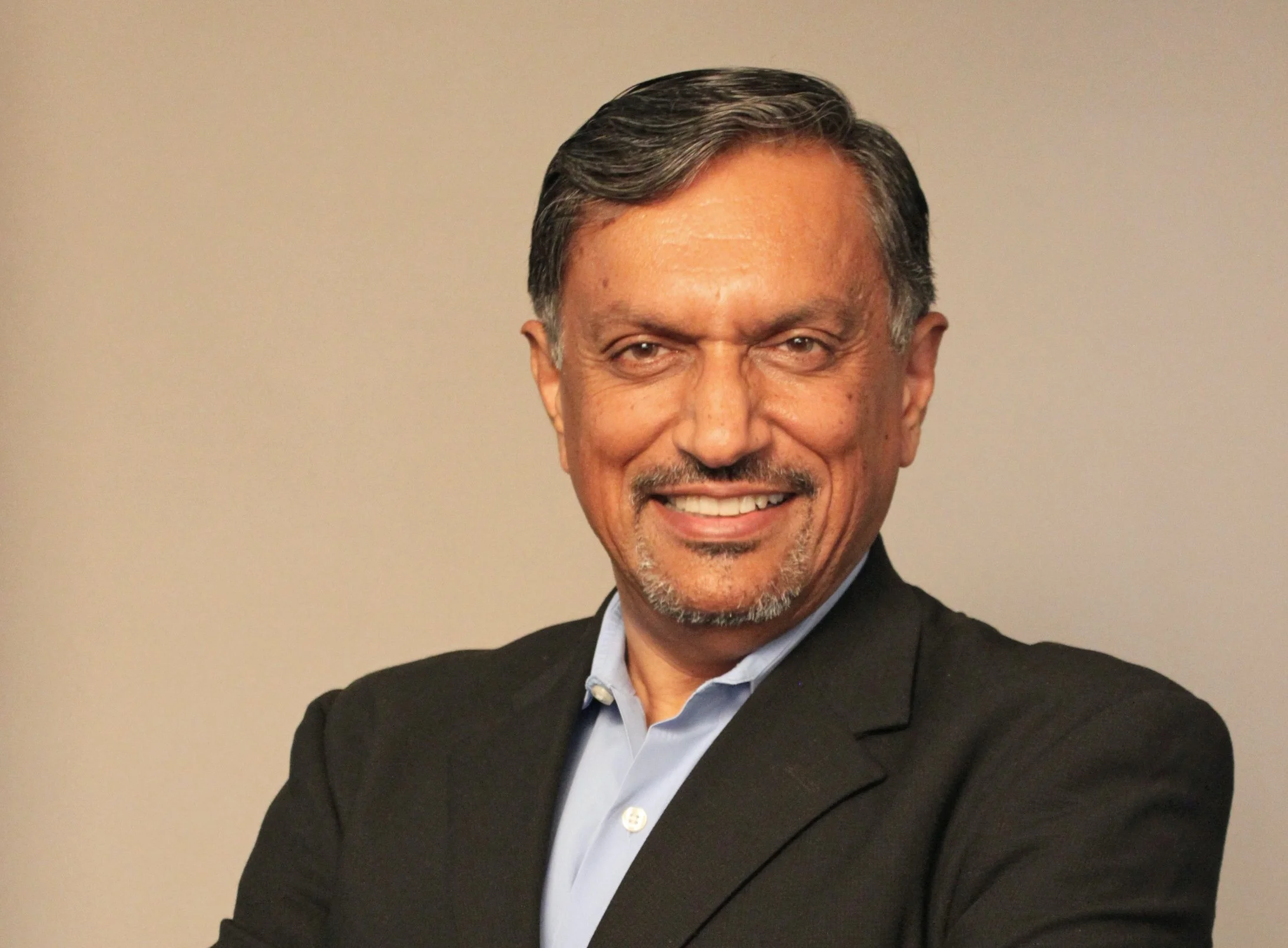 A middle-aged man with gray hair and a short beard, smiling, dressed in a black blazer and light blue collared shirt, against a plain beige background.