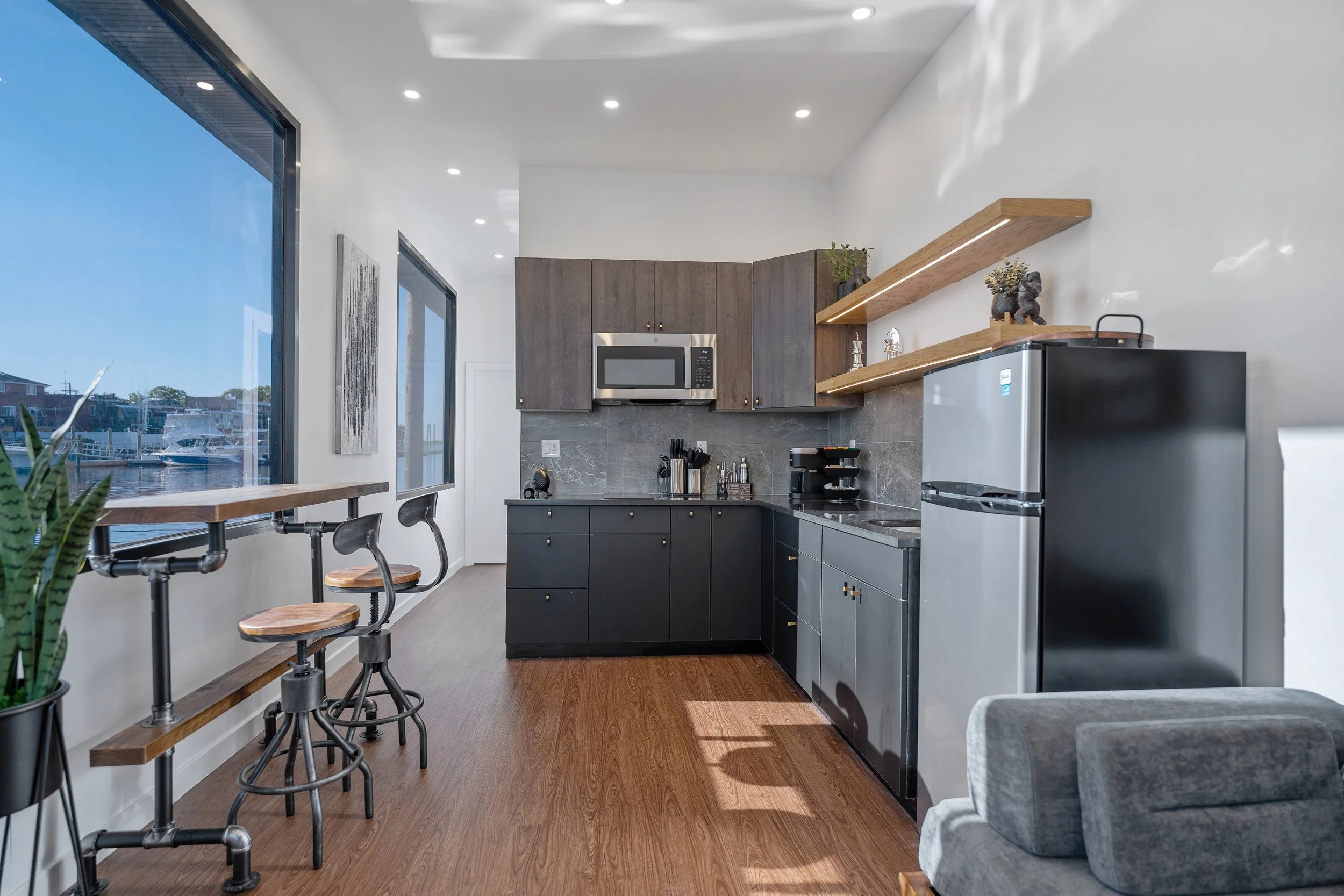 Kitchen with wooden floors, white walls, large windows overlooking a marina, black lower cabinets, brown upper cabinets, stainless steel microwave, small fridge, open shelves with decorative items, kitchen countertop with utensils, and a gray couch nearby.