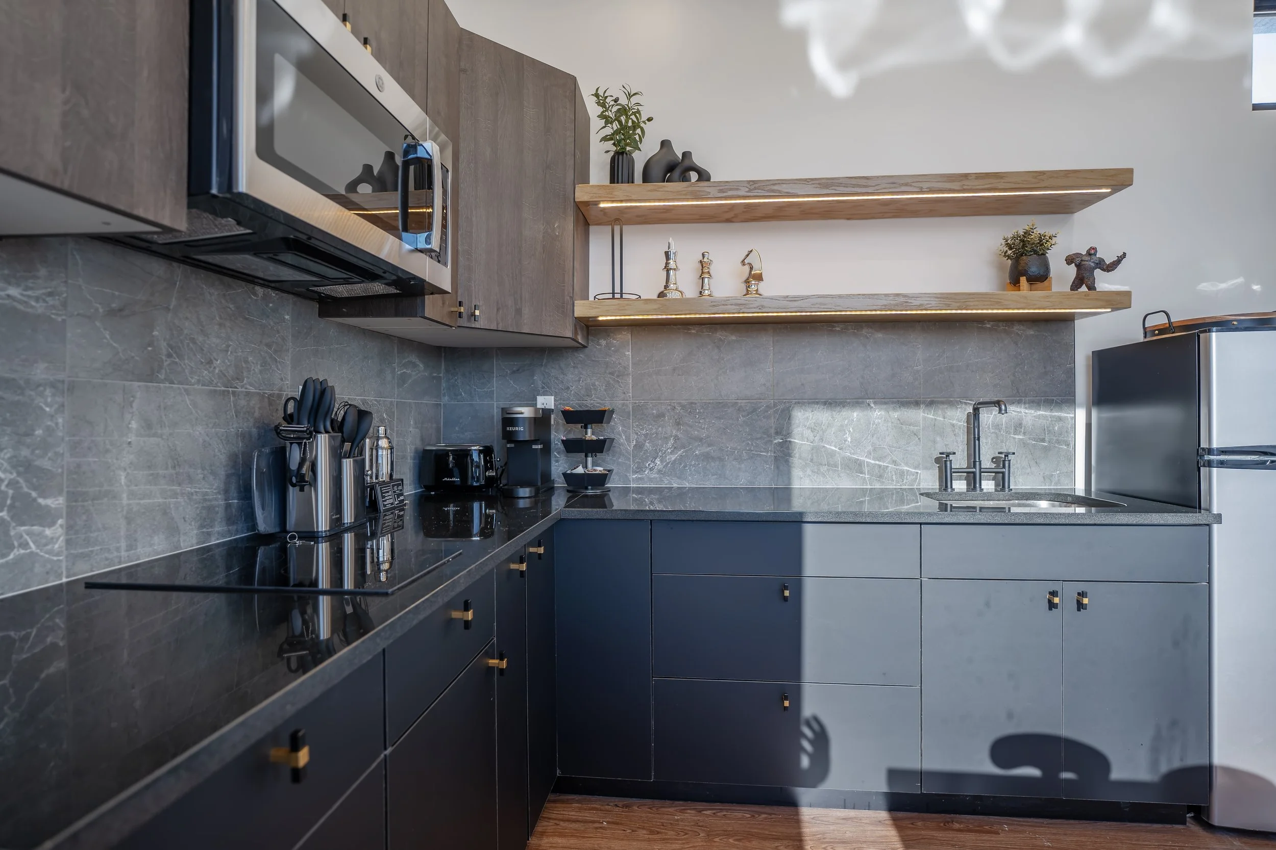 Modern kitchen with dark cabinets, a black marble countertop, and stainless steel appliances. Open wooden shelves with decorative items and plants, a sink, and a refrigerator are also visible.