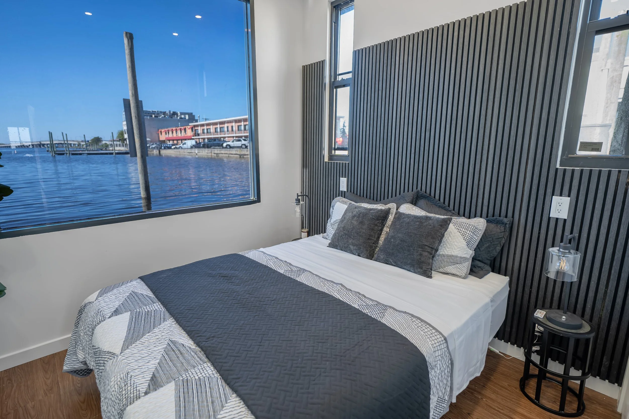 Bedroom with a bed, pillows, and a large window showing a view of a marina with boats and buildings, and a blue sky.