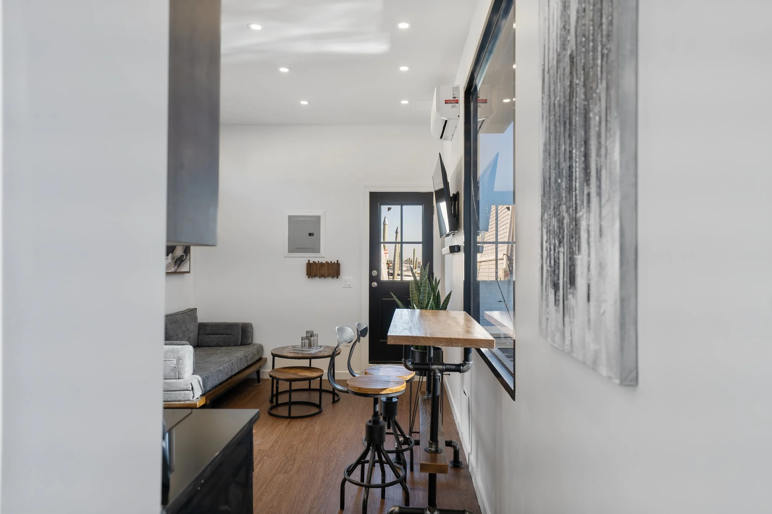 A modern, cozy living room with a gray sofa, round coffee tables, and a dining area with wooden tables and chairs. The space has white walls, wood flooring, and a black back door with a window. Recessed ceiling lights illuminate the room.