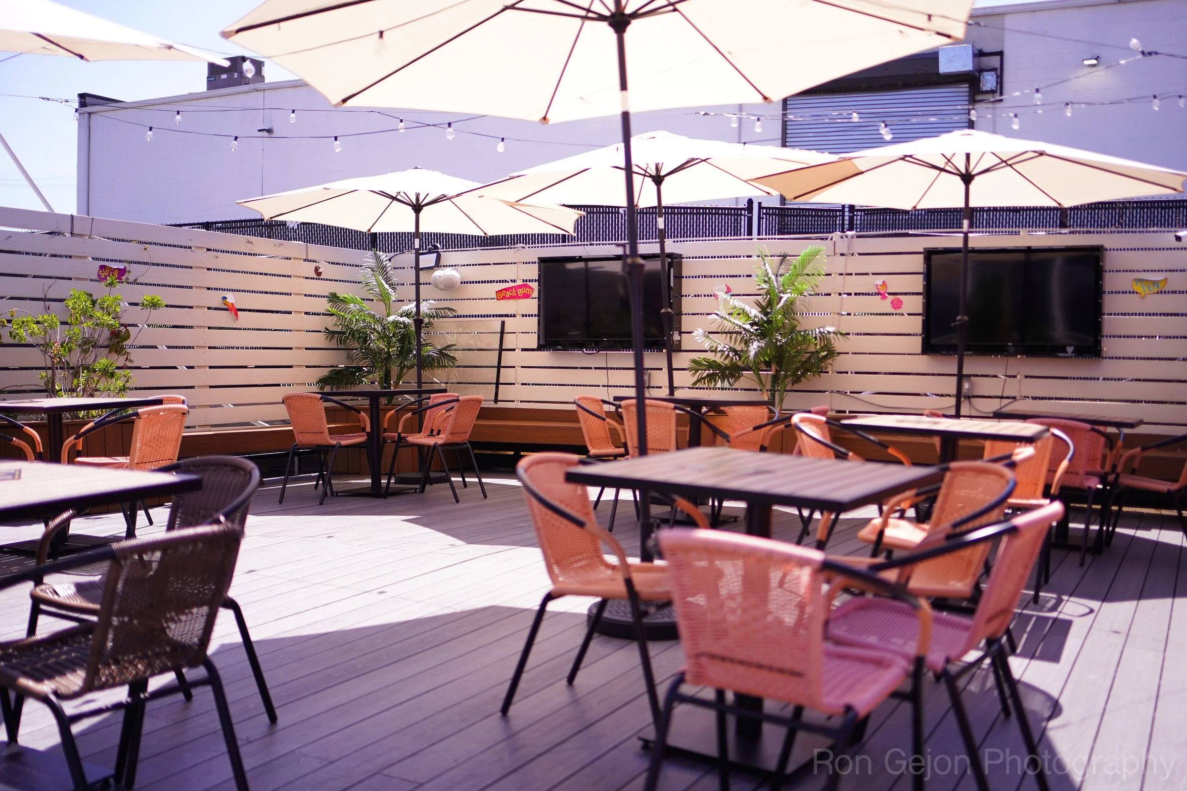 Outdoor rooftop bar with tables, chairs, large umbrellas, potted plants, string lights, and a sunset sky.