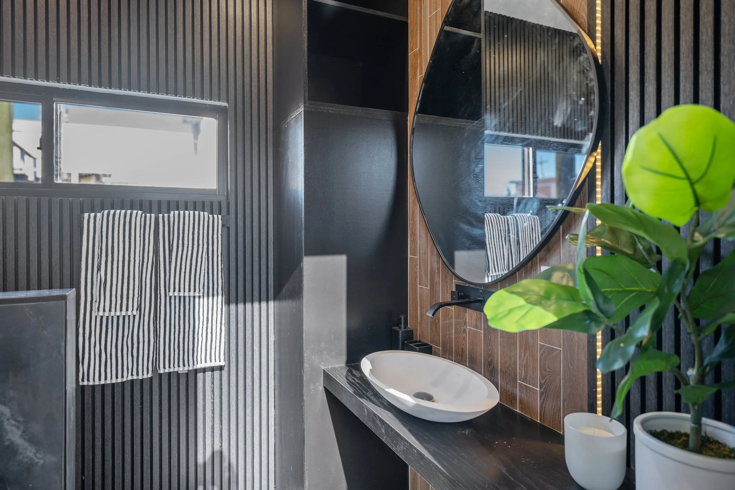 Modern bathroom with black textured wall panels, wooden accents, a large round mirror, a white bathtub, and potted green plants.