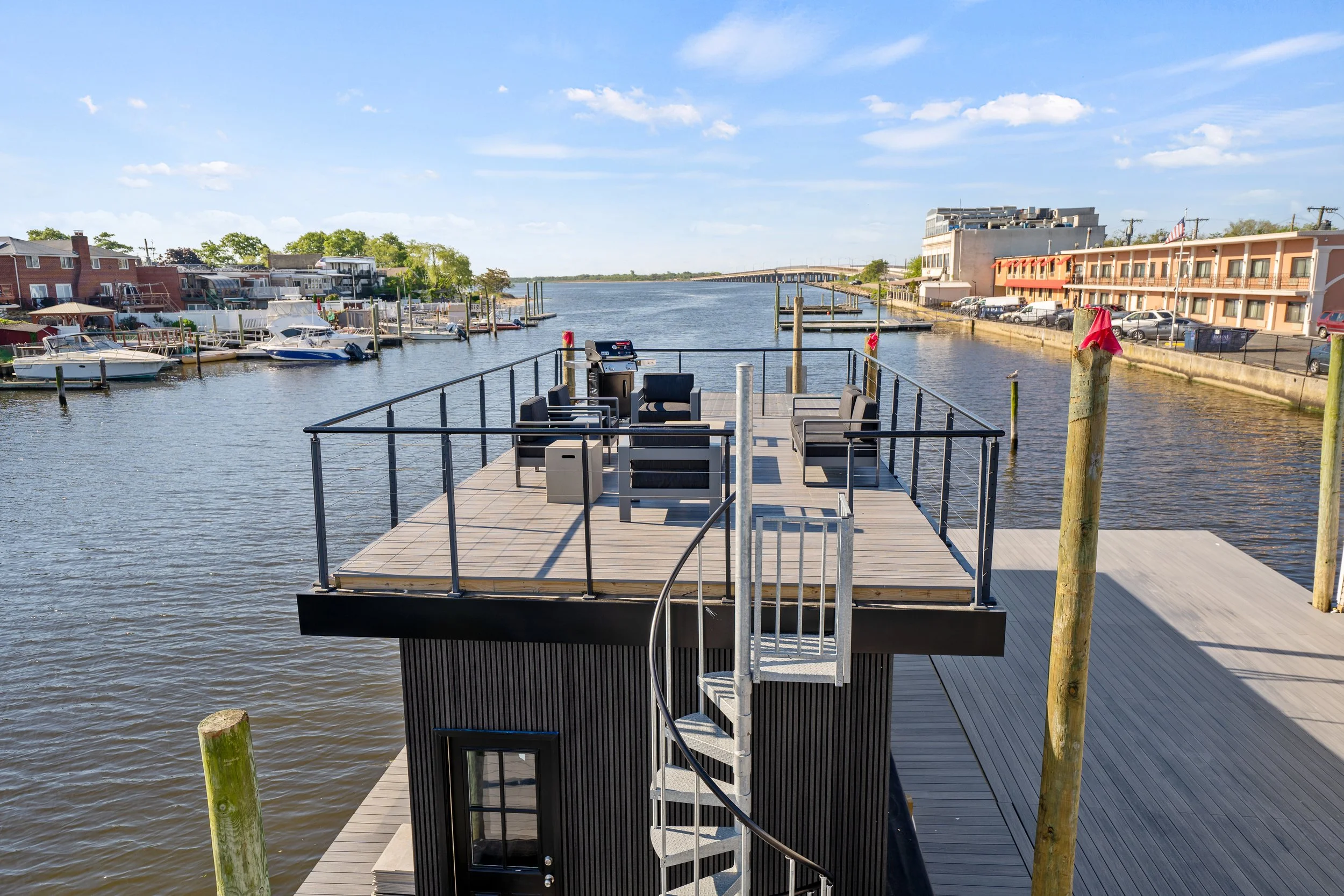 A waterfront view showing a floating deck with outdoor seating, boats moored along the dock, a bridge in the distance, and buildings lining the waterway under a partly cloudy sky.