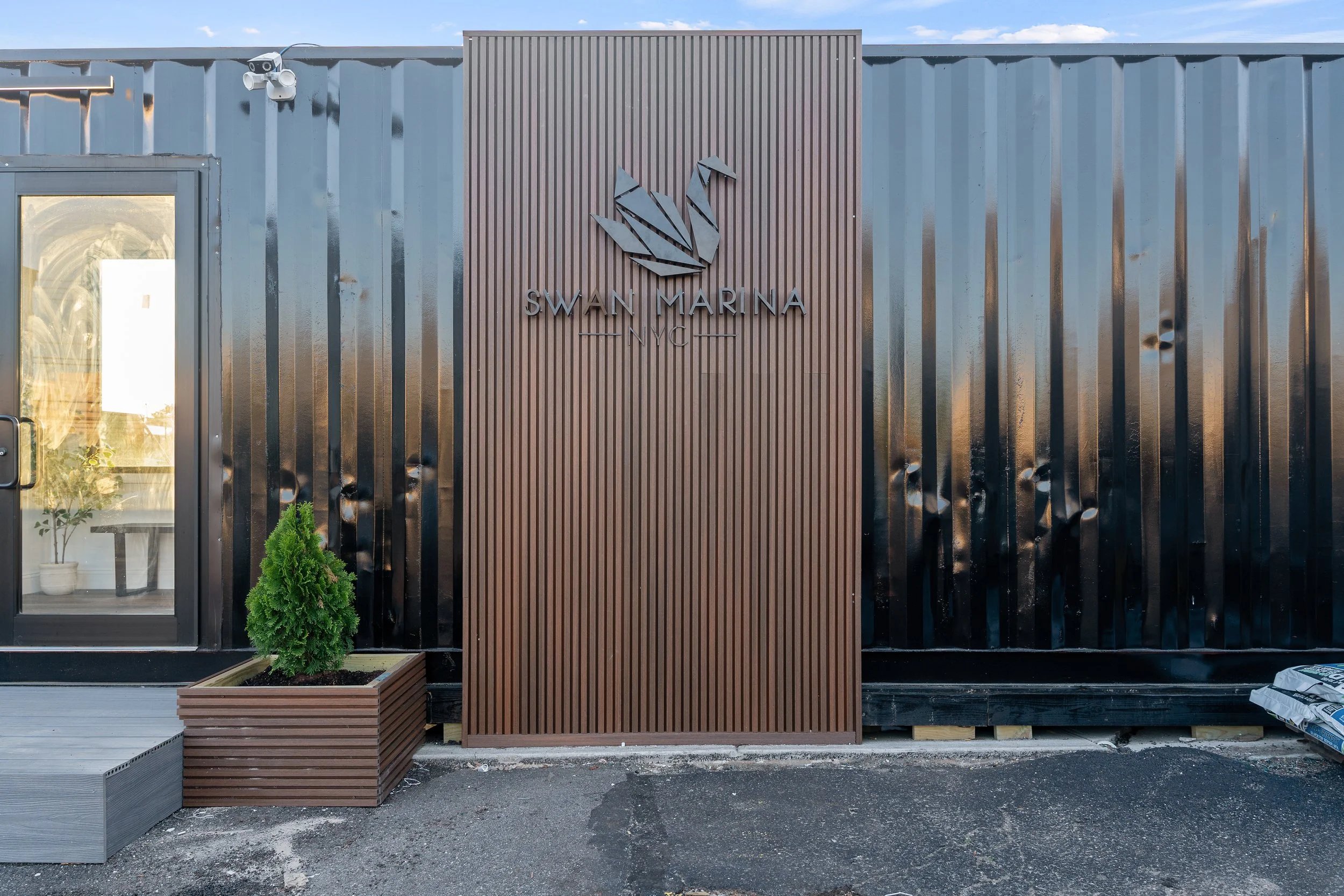 Exterior of a modern building with a sign reading 'Swan Marina Inc.' featuring a swan logo. The building has a dark ribbed metal facade with decorative wood paneling and a small potted plant in front.