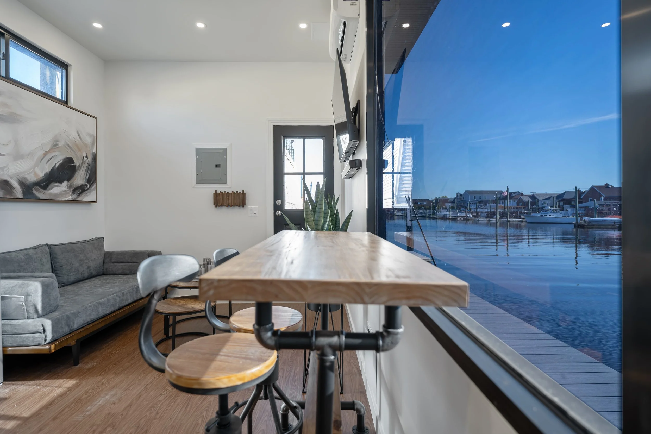 Interior of a modern living space with a gray sofa, abstract wall art, a wooden table, and black stools near large windows overlooking a marina with boats and waterfront houses.