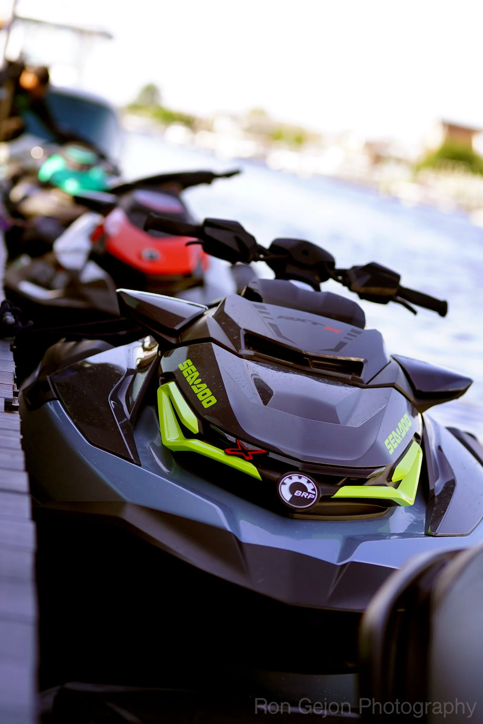 Multiple jet skis lined up on a wooden dock at a marina with boats and houses in the background under a partly cloudy sky.